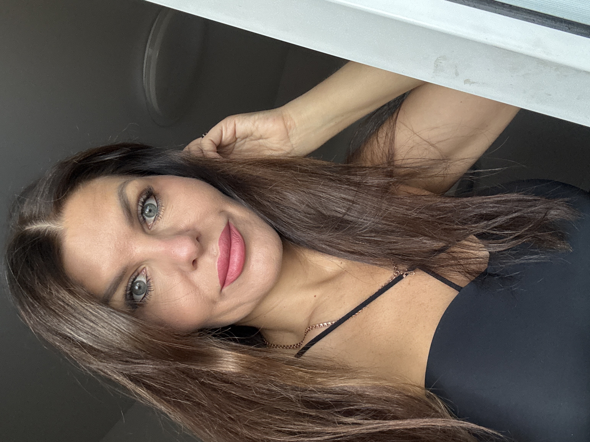 A close-up selfie of a woman with long, dark brown hair and light blue eyes. She is wearing a black top and a delicate gold chain necklace with a cross pendant. She is touching her hair with her right hand, and her left hand is not visible. The background is dark and out of focus, with a sliver of a white structure on the right side of the image. The image does not provide any information about the city of Chaikovsky, Russia.
