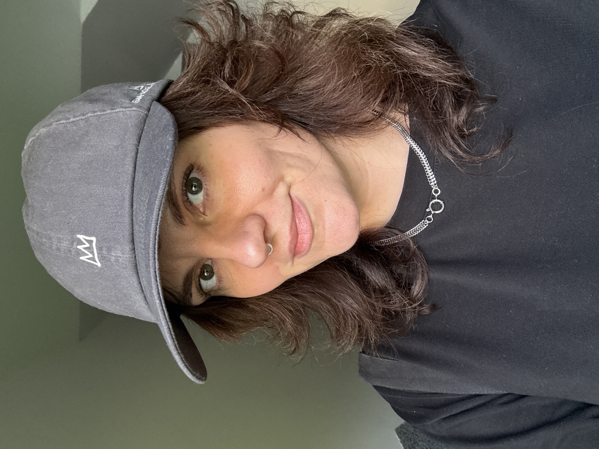 A woman is seen from a low angle in a close-up shot. She has brown, wavy hair that is shoulder-length, wearing a grey baseball cap with a white crown logo. She is wearing a black shirt and a silver chain necklace. She has a nose ring on her left nostril. The background is a light, solid color. There are no visible details of Boreti, Montenegro, in the image.