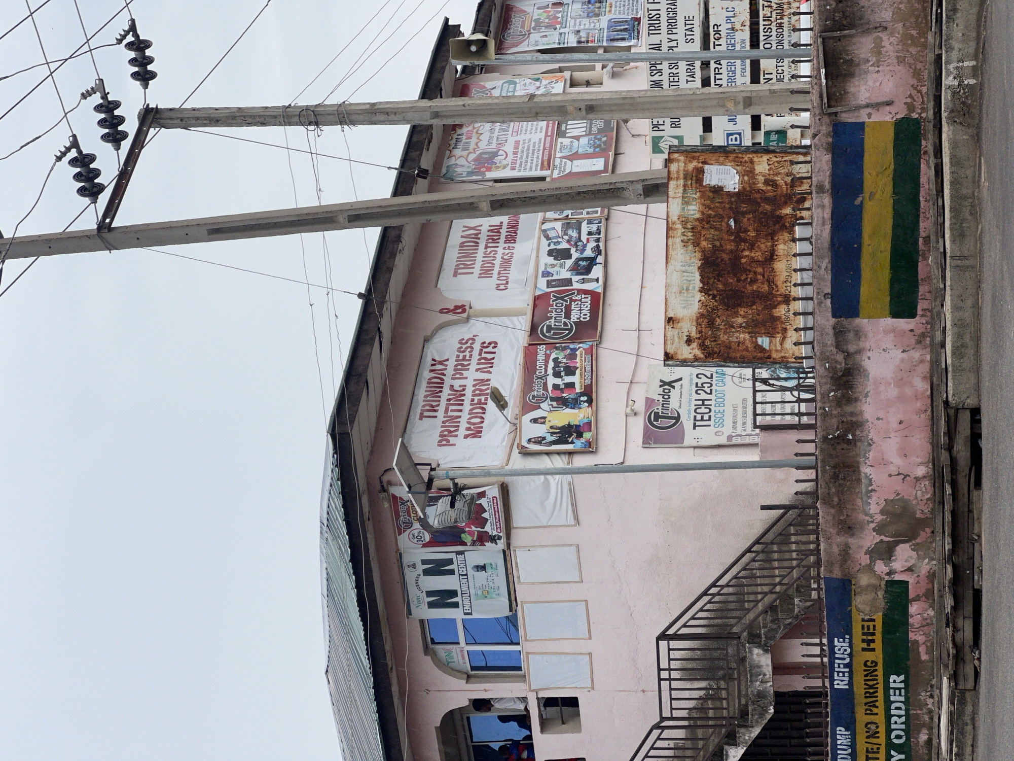 The image shows a two-story building with a light pink facade, located on a street in Jalingo, Nigeria. The building has a metal corrugated roof and is adorned with various signs and advertisements. On the first floor, there is a prominent sign for "N NAME LICENSED ENROLLMENT CENTRE" featuring a portrait of a man. Other signs advertise "TRINIDAX PRINTING PRESS, MODERN ARTS" and "TRINIDAX INDUSTRIAL CLOTHINGS & BRANDING." Several windows, some with blue tinted glass, are visible on the facade. A set of metal stairs with railings leads up to an entrance on the left side of the building.
In front of the building, a concrete wall runs along the street, painted with horizontal stripes of blue, yellow, and green. A large, rusted metal panel is affixed to the wall, displaying "NIGERIAN..." and some illegible text. Another sign on the building, further to the right, advertises "JM SPECIAL TRUST FU..." and "TER SUPPLY PROGRAM."
Electrical poles with multiple insulators and wires are prominent in the scene, suggesting an urban or semi-urban environment. The street in the foreground is paved with asphalt and shows some signs of wear and debris. The overall atmosphere is overcast, with a pale sky. A few people are partially visible through the windows and at the entrance on the left. The scene depicts a commercial area with various services offered.