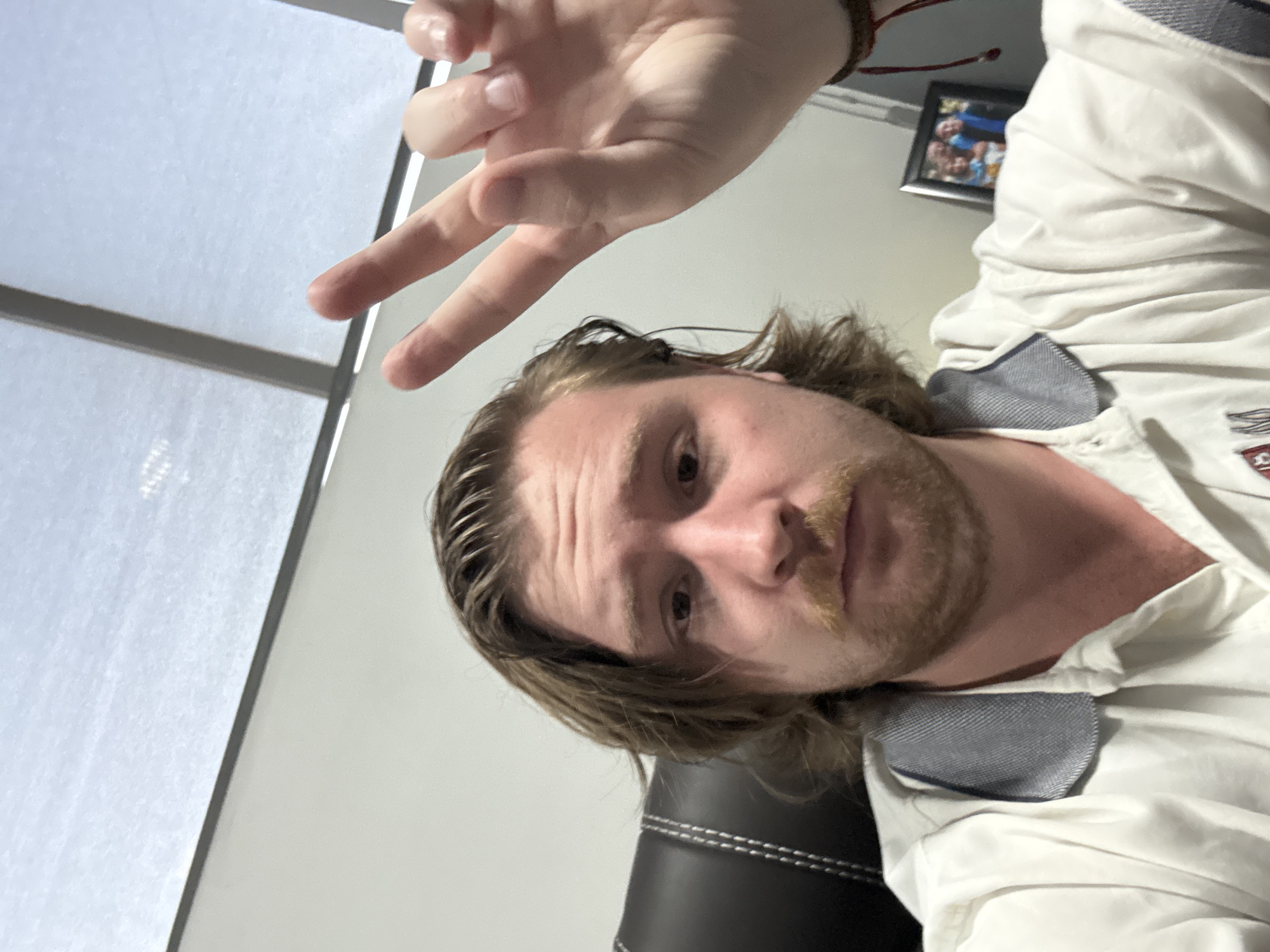 A man with shoulder-length blond hair and a mustache is facing the camera, making a "peace" sign with his right hand. He is wearing a white, collared shirt. The background consists of a window with blinds, a white wall, a picture frame, and a black chair. The location is presumably indoors in Cuautitlán Izcalli, Mexico.