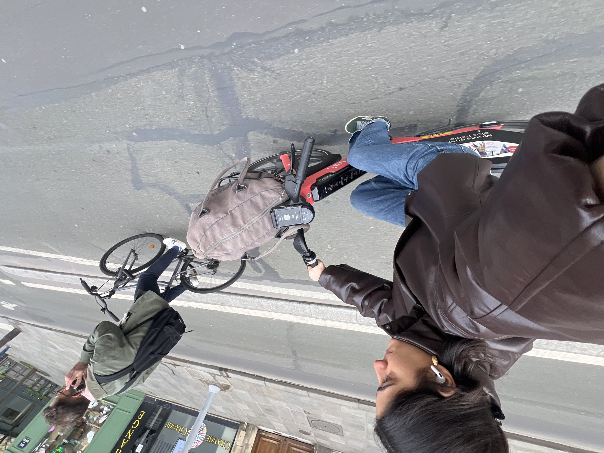 Here is a detailed description of the image:
A street-level, eye-level shot depicts two individuals in an urban setting, likely Paris, France. The photograph appears to be taken from a slightly rotated perspective.
In the foreground, a person in a brown leather jacket and jeans rides a red e-bike, possibly a "Volta" rental bike, based on visible branding. The individual is holding a phone attached to the handlebars.
Behind, there is another person riding a bicycle. They wear an olive-green jacket and dark trousers, along with white sneakers. A black backpack is slung across their back.
The background features the urban landscape with sidewalks and the corner of a building displaying the word "CHANGE".