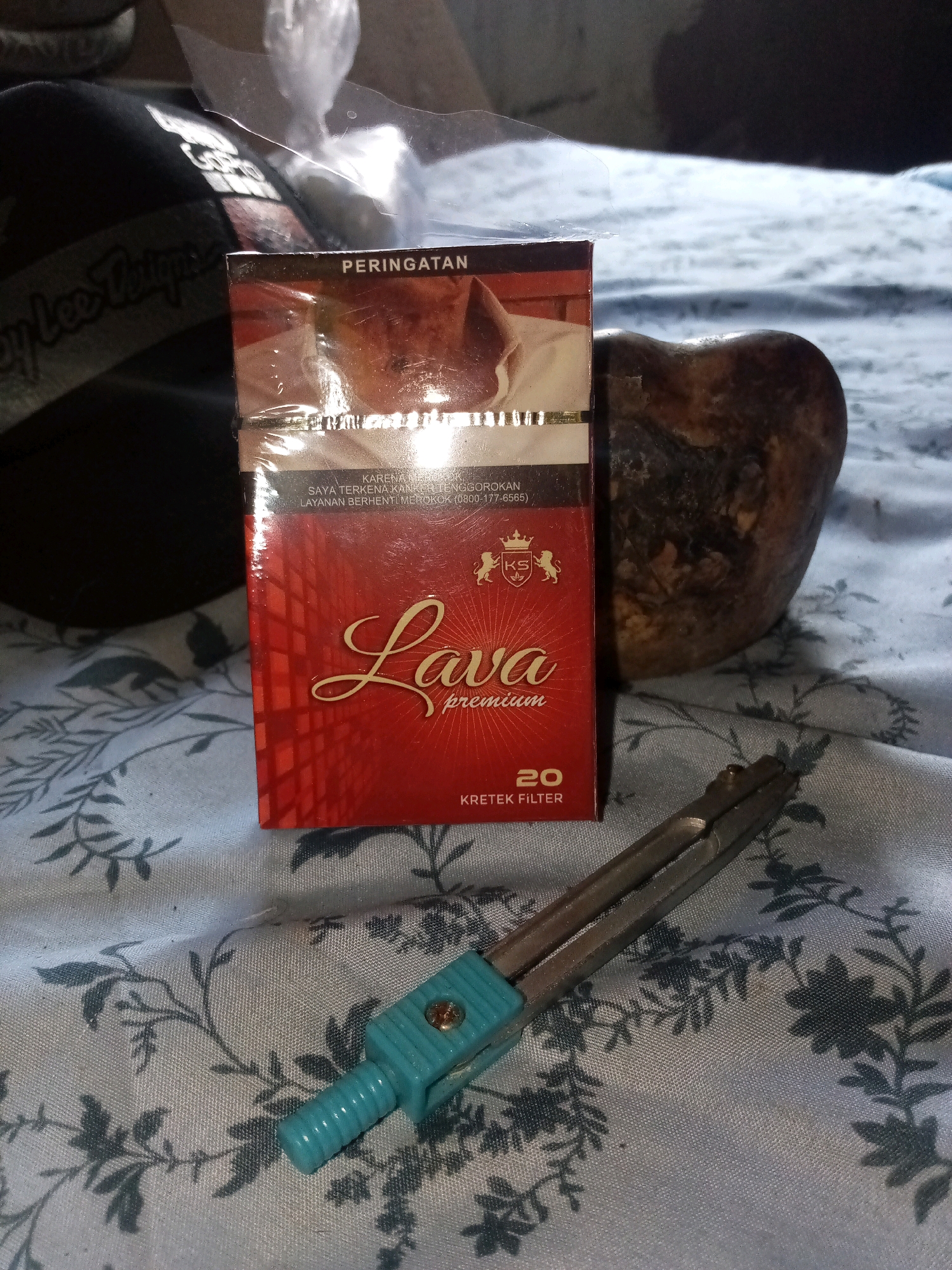The image shows a close-up, eye-level view of a pack of "Lava premium" kretek filter cigarettes, lying on a patterned cloth. To the left of the cigarette pack is a dark baseball cap with lettering that appears to read "Lee Thipe". To the right of the pack is a brown, irregular-shaped object, possibly a rock or a piece of wood. In the foreground, a teal and silver drawing compass is positioned diagonally across the cloth. The background is blurry but appears to be a flat surface. The lighting suggests an indoor environment.