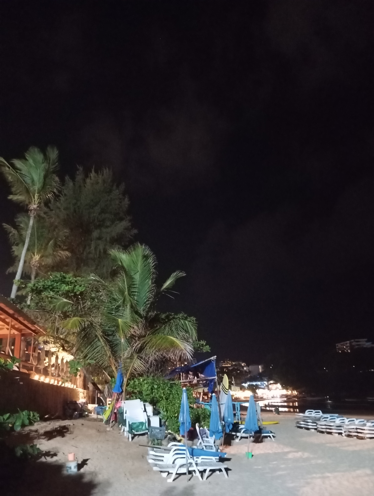 This image captures a nighttime scene on a beach in Karon, Thailand, with a focus on the sandy shore and surrounding vegetation. The sky is dark, indicating it is likely past sunset, with a few faint clouds visible.
In the foreground, the sandy beach is sparsely populated. Several rows of empty lounge chairs, predominantly white and made of plastic, are stacked or laid out, suggesting preparation for daytime use. Blue beach umbrellas, some open and some closed, are scattered amongst the chairs.
To the left of the frame, a building with a sloped, orange-tinted roof is partially visible. It appears to be a restaurant or bar, illuminated by warm, string lights that cast a glow along its facade. Green foliage, including palm trees with their characteristic fronds, surrounds the building and extends towards the beach. Some of the palm trees are tall and slender, while others are more mature with wider canopies.
In the background, across the water, a distant city skyline is visible, characterized by rows of lights indicating buildings and urban development. The water itself is dark, reflecting the night sky and the lights from the distant shore. The overall atmosphere is one of a quiet, nocturnal beach setting, transitioning from day to night.