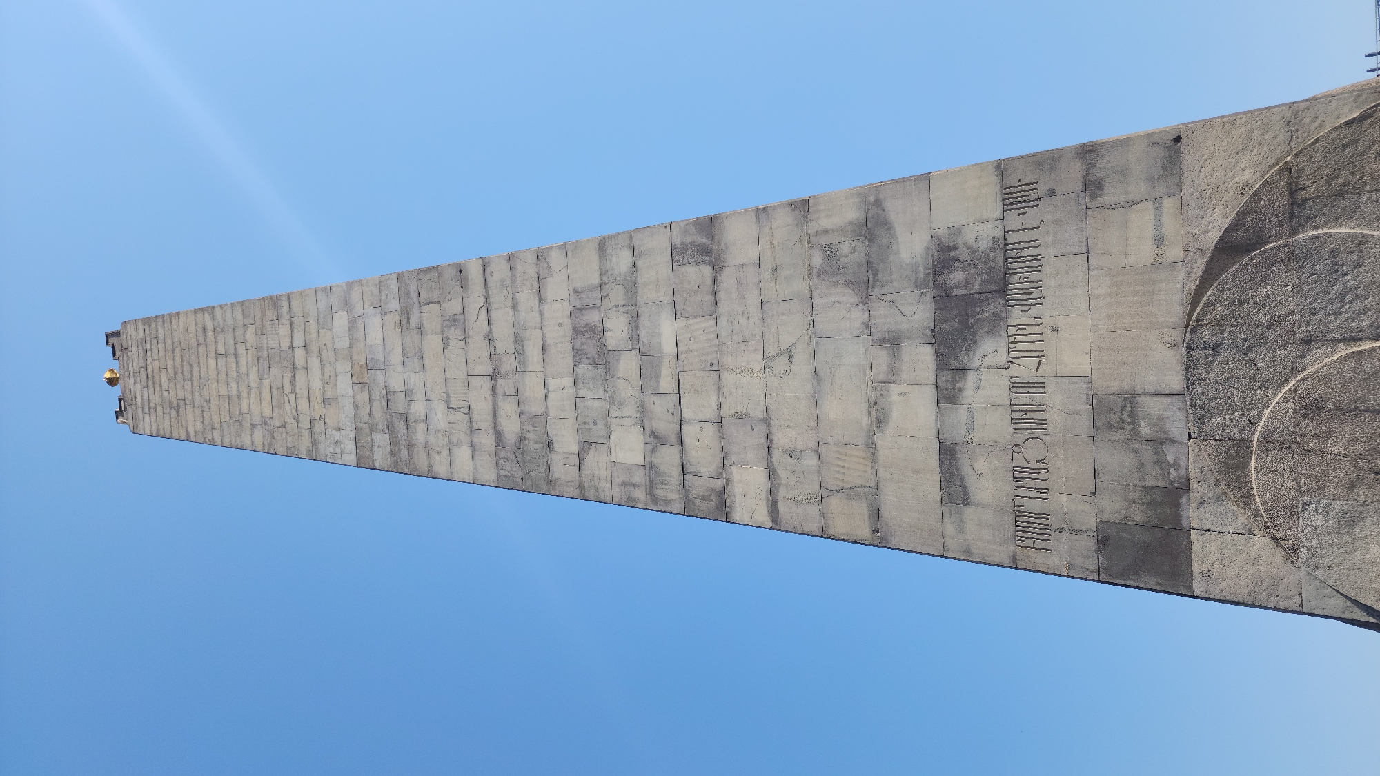 A low-angle, full shot captures a towering, trapezoidal stone monument against a clear, bright blue sky. The monument is constructed from rectangular stone blocks, some of which exhibit staining and wear, contributing to a weathered appearance. Inscriptions are carved into the stone in an unfamiliar script. Near the base, a circular, textured relief is visible. The monument tapers upwards, culminating in a flat top with crenellated features and a golden orb or finial. The bright sunlight casts minimal shadows, indicating a midday or near-midday sun. The monument is situated in Yerevan, Armenia, specifically at the Tsitsernakaberd memorial complex, a poignant site dedicated to the victims of the Armenian Genocide. No people are visible in the frame.
