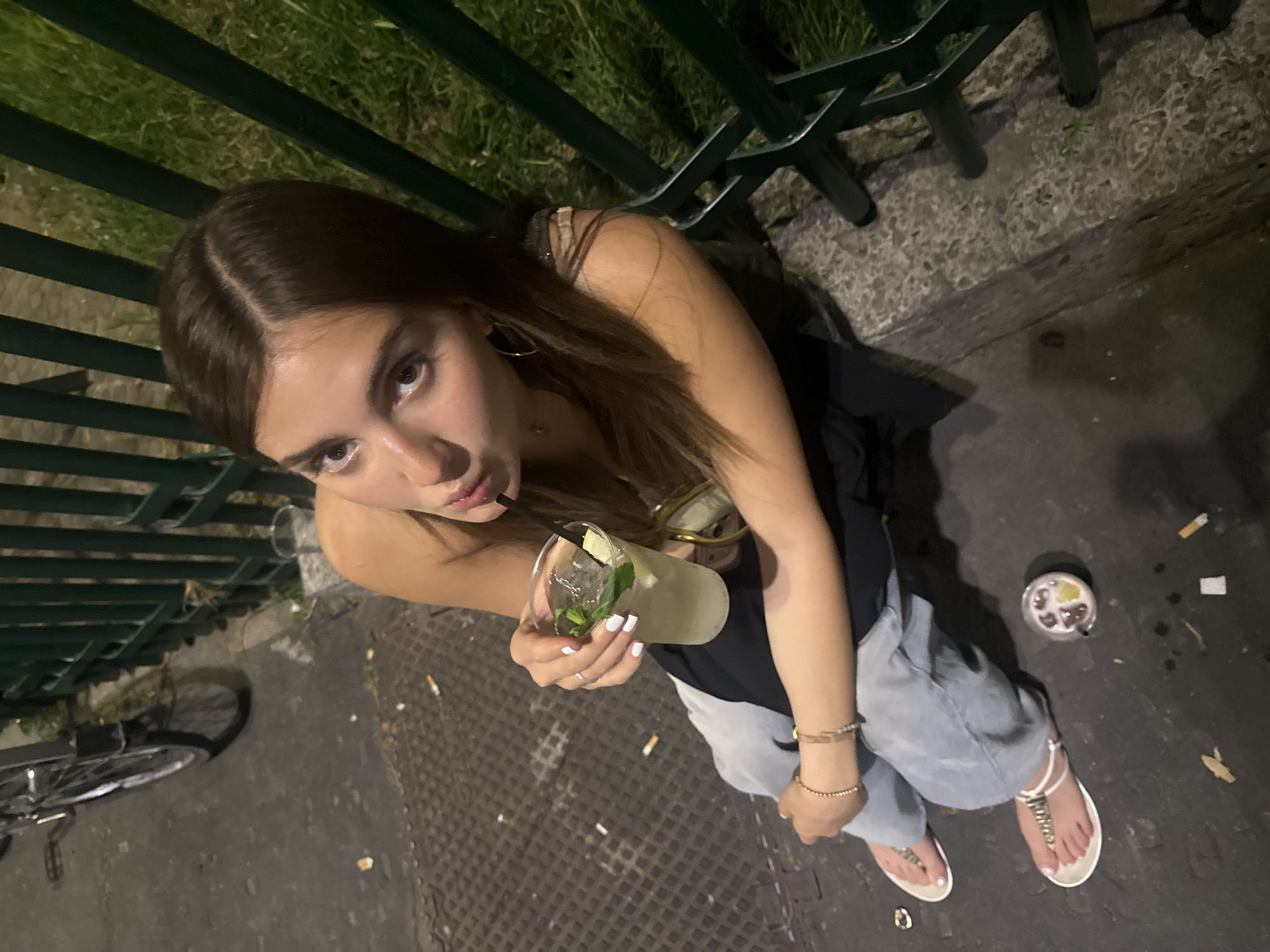 A high-angle, close-up photograph shows a young woman holding a drink. She has long, light brown hair and is wearing hoop earrings, a black top, and light-wash jeans. She is standing in a paved area scattered with cigarette butts and discarded objects, including a plastic cup with remnants of a drink. She is holding a clear plastic cup with a beverage and a black straw. The drink contains ice, mint, and a slice of lemon or lime. She is wearing white sandals with a T-strap design. A section of a green metal fence is visible in the upper portion of the frame. A bicycle wheel is partially visible on the left side of the frame. The location appears to be an urban outdoor setting.