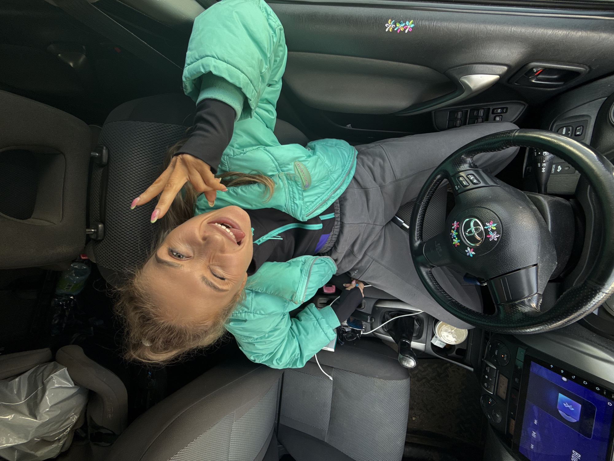 The image presents a playful scene inside a car, likely in Chaikovsky, Russia, captured from a top-down perspective. A woman with light hair, styled in a bun, is lying across the front driver's and passenger seats. She is wearing a bright turquoise puffy jacket over a black long-sleeved top and gray pants. Her face, seen upside down from the camera's angle, displays a wide, joyful smile with her tongue slightly out and eyes happily crinkled. She holds up her right hand, featuring nails with pink and green gradient tips, in a V-sign gesture towards the camera.
The car's interior features prominently, including the dashboard, steering wheel, and door panels. The steering wheel bears a "TOYOTA" logo adorned with colorful flower stickers, and similar flower decals are visible on the car's door panel. A large, modern media screen is installed in the dashboard, displaying a "Bluetooth" audio interface. Other visible objects include car controls, a gear stick, charging cables, sunglasses, a water bottle, and a plastic bag on the passenger-side floor. The woman's jacket has a subtle "Marmot" logo visible. The setting suggests a casual, possibly travel-related moment. The overall atmosphere conveyed by the woman's expression and pose is one of happiness and lightheartedness.