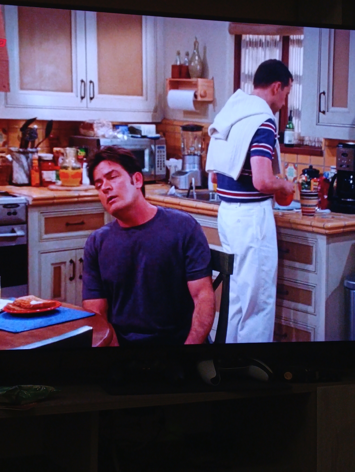 The image shows a scene from a television show, likely a sitcom, playing on a TV screen. Two men are visible in a kitchen setting. The man on the left is seated at a table with his head resting on his shoulder. He is wearing a dark purple t-shirt. On the table in front of him is a plate with food on it. The man on the right is standing at the kitchen counter. He is wearing a blue and white striped shirt with a white sweater draped over his shoulders, and white pants. He is holding an orange in his hands. The kitchen has light-colored cabinets and a patterned backsplash. A window with white curtains is visible in the background. The TV screen is resting on a shelf, and a game console controller is visible below the screen.
There is no evidence to suggest that this image or scene is in Plovdiv, Bulgaria. It is a generic kitchen scene that could be from any television studio.