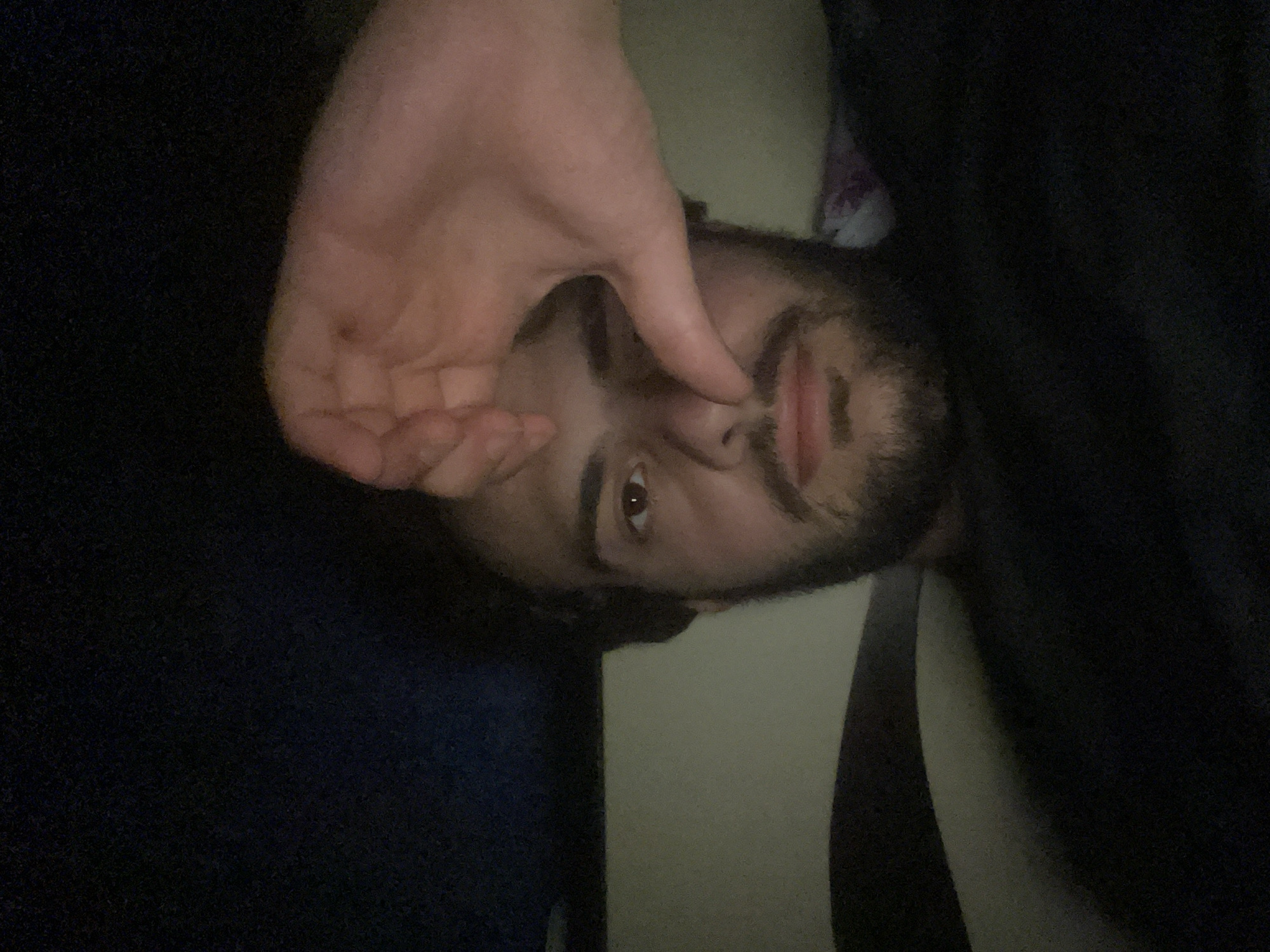 The image is a selfie taken in a dimly lit room, possibly a bedroom. The subject is a young man with dark hair and a beard, looking at the camera. He is wearing a dark shirt. His right hand is raised, with his thumb and index finger forming a shape over his nose and eye. The background is dark, with a hint of a lighter surface behind him and some colorful fabric to the right. The image does not contain any information that would allow for the identification of its location in Tbilisi, Georgia.