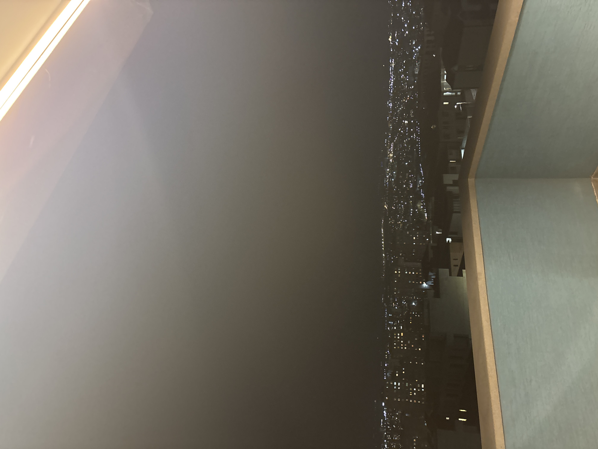The image displays a panoramic nighttime view of a city, likely taken from a high vantage point such as an apartment window or balcony. The dominant features include a bright, elongated linear light source, possibly an interior light fixture or a window frame, in the top-left quadrant, casting a soft glow across the upper portion of the frame. The central area of the image is dominated by a dark, hazy expanse, suggesting either a foggy or misty night, or significant air pollution.
To the right, a sprawling urban landscape unfolds, characterized by numerous twinkling lights emanating from buildings and streets, stretching horizontally across the scene. These lights, though somewhat obscured by the atmospheric haze, indicate a dense residential or commercial area. On the far right edge, a distinct architectural element is visible – a light blue or green wall or railing with a beige or tan upper edge, suggesting part of the interior space or a balcony from which the photograph was taken.
The overall scene is a static, wide shot of an urban environment at night. No people or specific activities are visible. The time of day is clearly nighttime due to the artificial lights illuminating the city. The pervasive haze contributes to a somewhat blurred and atmospheric quality. There is no visible text. Given the context, this is a nighttime view of Arinj, Armenia.