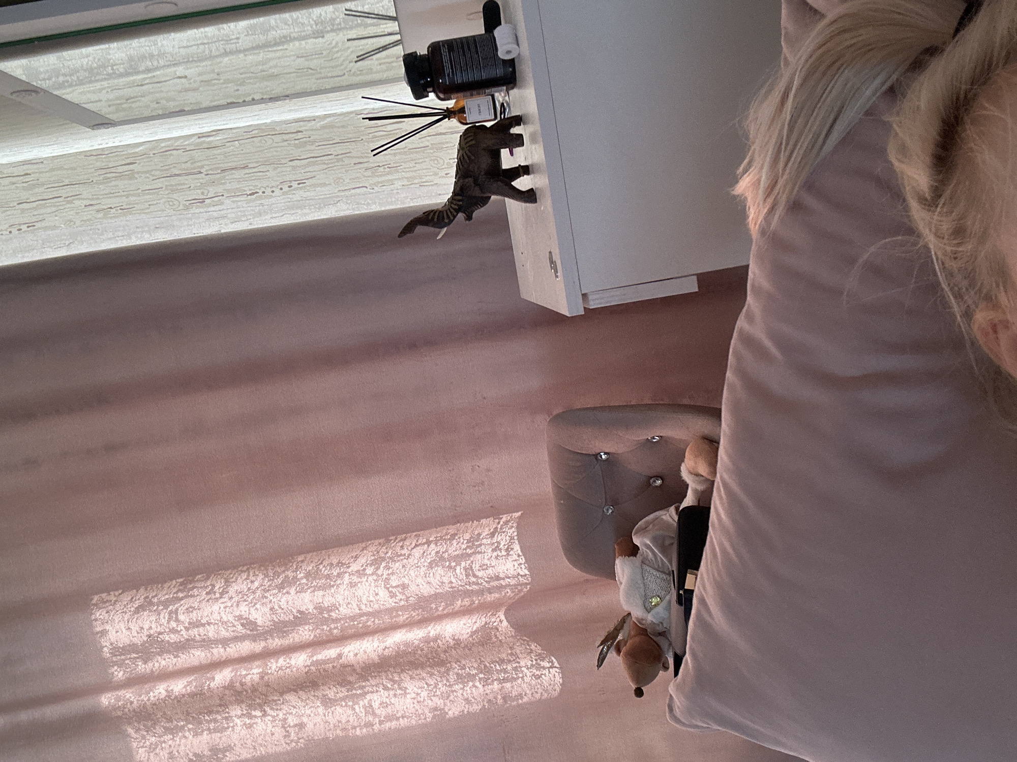 The image depicts an indoor scene, rotated such that the left side appears as the top. In the upper portion of the image, a white bedside table is visible, with a diffuser and a dinosaur toy placed on top. The background features a window or reflective surface.
In the lower half, there is a pink carpet, a pink headboard with decorative buttons, and a stuffed animal sitting on the headboard. The angle suggests a perspective from a high vantage point, possibly above a bed. The scene is situated in a room, with lighting and furnishings. No location information for the city of Высокий, Russia, is discernible from the visual content of this image.