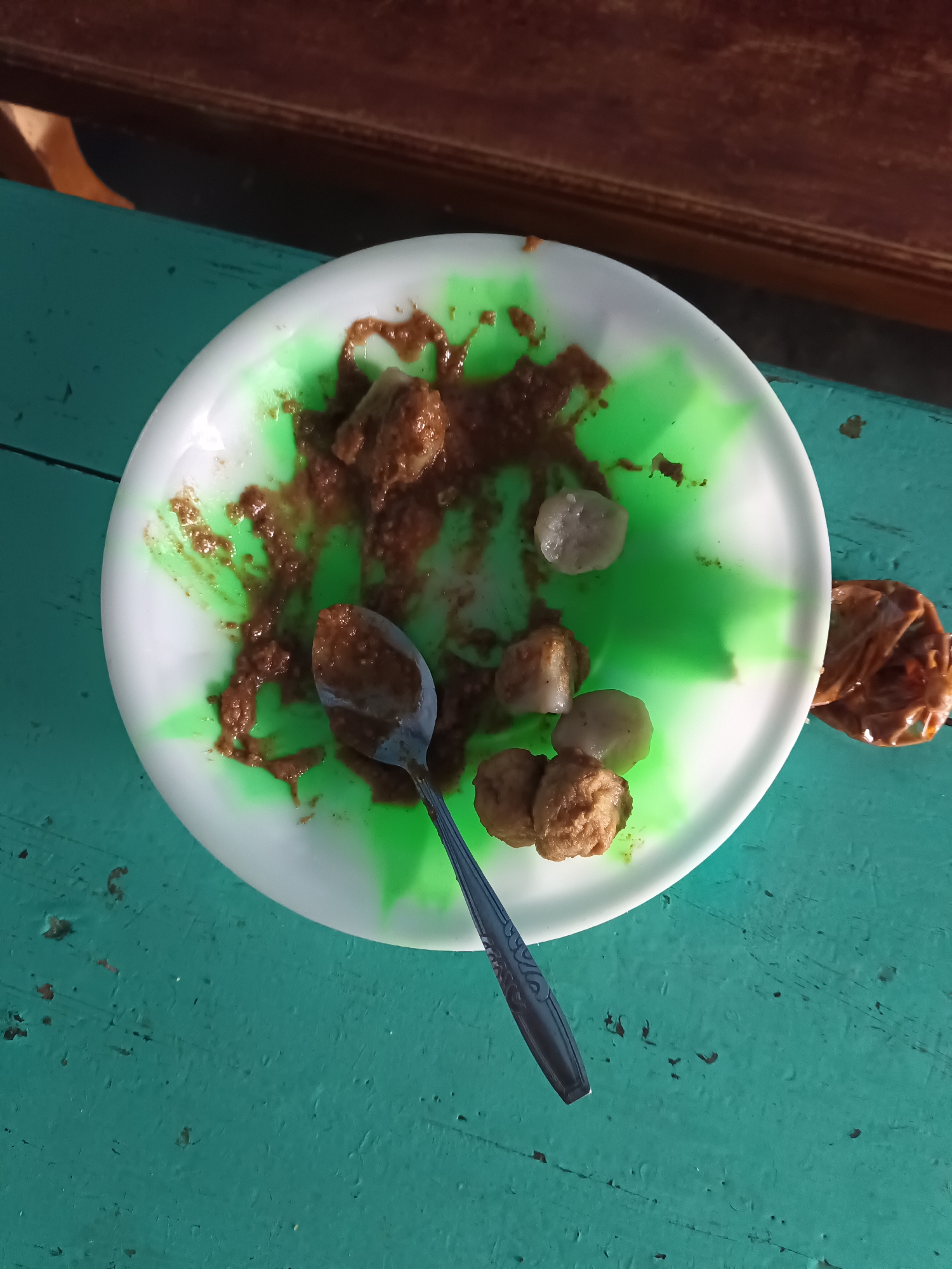 The image shows a close-up, top-down view of a white plate with a green swirl design, containing a few meatballs and a dollop of brown sauce. A silver spoon rests on the plate, also coated in sauce. To the right of the plate, a crumpled brown paper bag with a similar sauce is visible. The plate and bag are positioned on a teal-colored wooden surface that appears to be outdoors, possibly a table. The background is blurred, with hints of a wooden structure and sunlight. The scene appears to be an outdoor meal, likely in a casual setting. There is no specific indication of the location being in Terbanggi Marga, Indonesia, beyond the user's prompt.