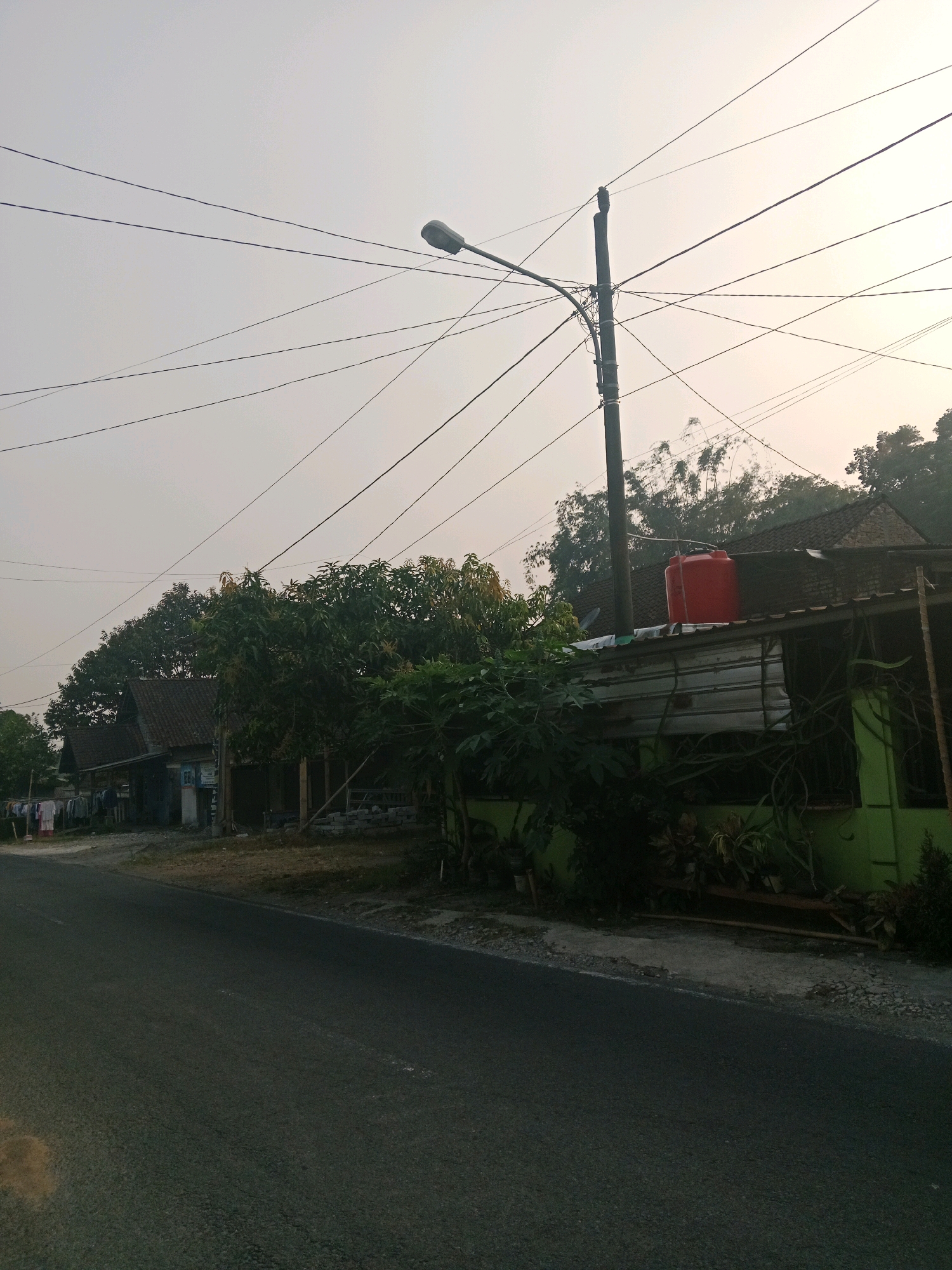 The image captures a street scene in Salatiga, Indonesia, under an overcast sky. In the foreground, a paved road occupies the lower half of the frame. To the right of the road, a low-lying green building is visible, partially obscured by lush green foliage. A large red water tank sits atop the roof of this building. A prominent utility pole, laden with numerous power lines that crisscross the sky, stands near the building. Beyond the green structure and utility pole, more houses with tiled roofs and trees are discernible. To the left of the road, further houses are present, with laundry hanging out to dry in front of one of them. The overall impression is of a residential area on a hazy day.