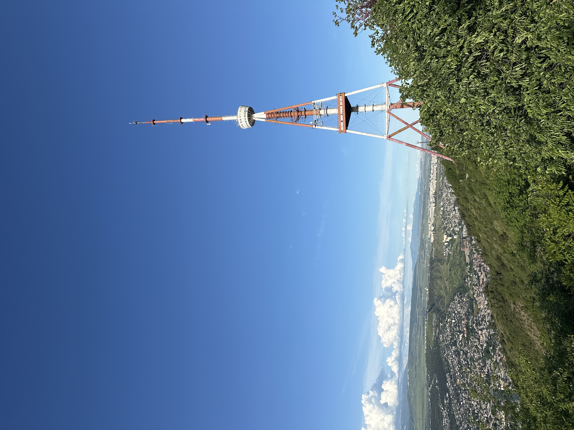 The image shows the Tbilisi TV Broadcasting Tower, a prominent structure in Tbilisi, Georgia. The tower is painted in alternating red and white sections and stands against a clear blue sky. Part of a city is visible in the background, along with green hills and vegetation. Some trees obscure the base of the tower. The landscape extends to a visible horizon with white clouds.