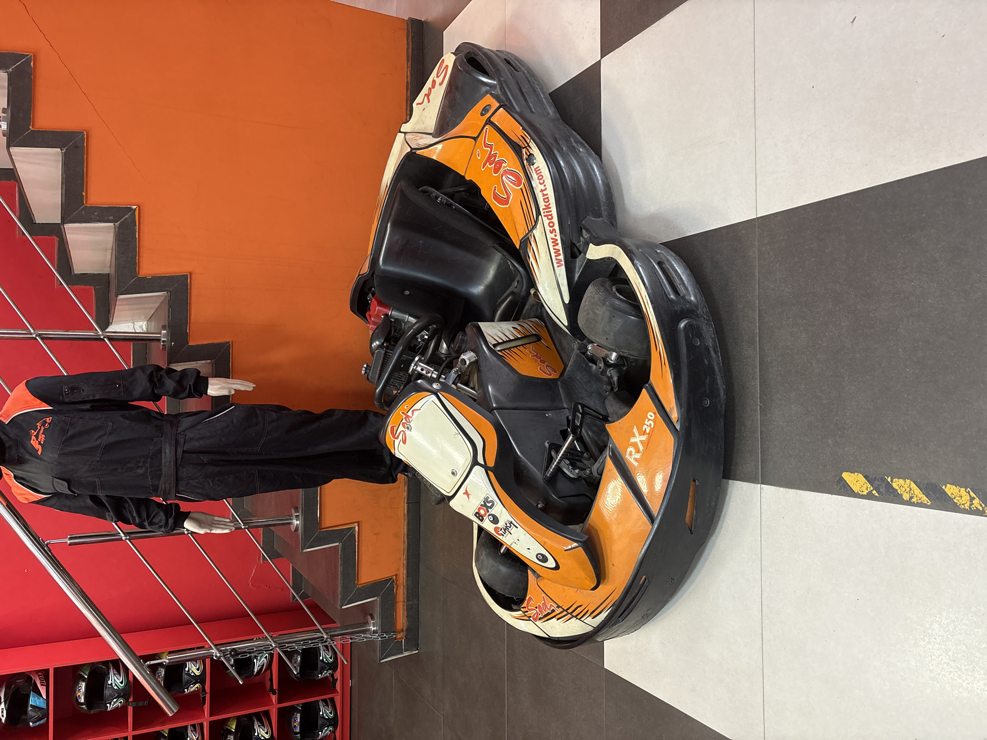 In Saint Petersburg, Russia, a go-kart track facility is depicted. A single go-kart, predominantly orange and white with black accents, rests on a black and white checkered floor. The go-kart features the branding "Sodikart" and "RX 250." Behind the go-kart, a set of stairs with red walls and orange risers ascends. A mannequin, dressed in a black and orange racing suit, stands at the top of the stairs, facing forward. To the left of the stairs, shelves are visible, containing several racing helmets. The overall environment suggests an indoor recreational space dedicated to go-karting.