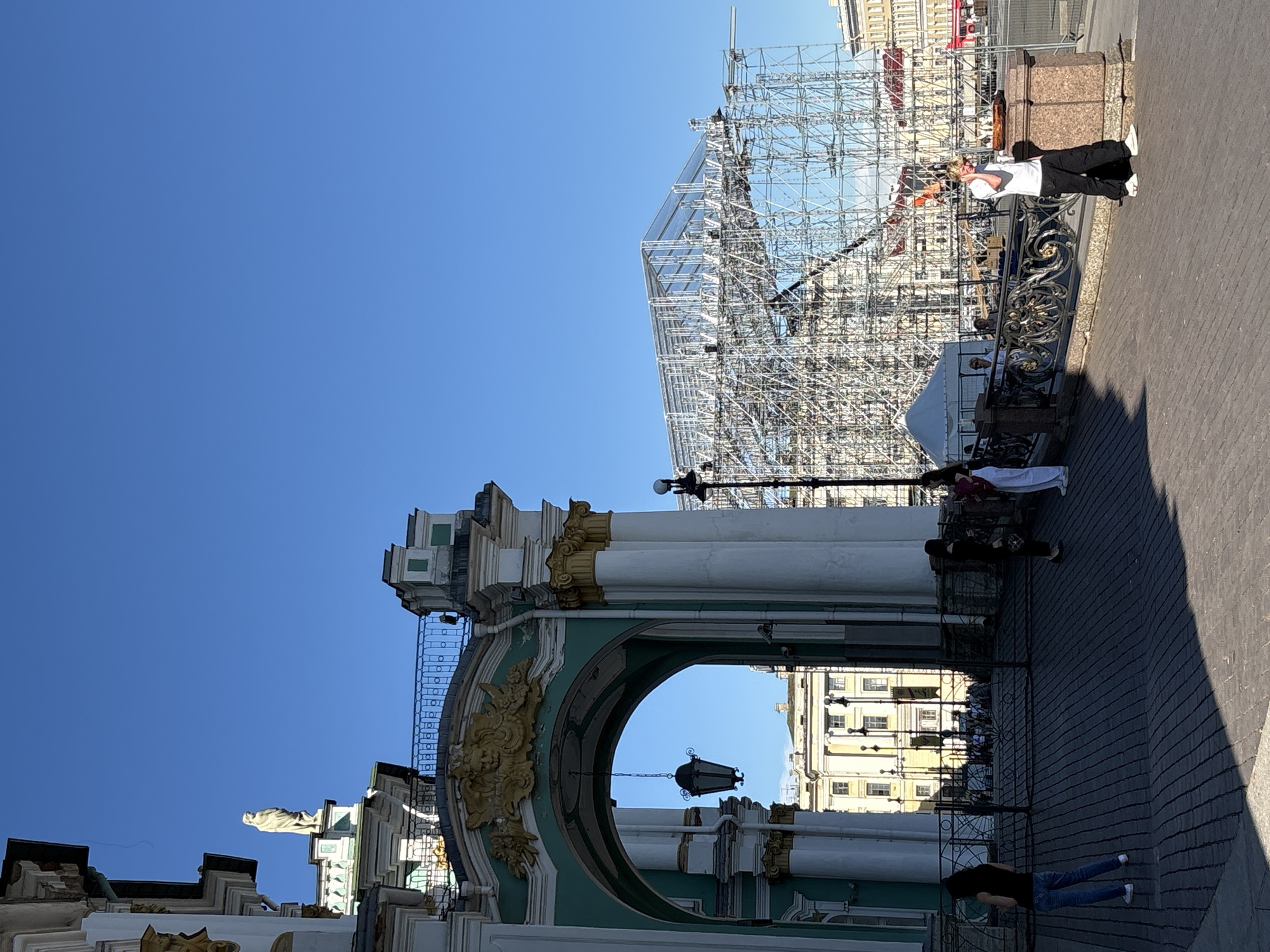 The image captures a sunny day at Palace Square in Saint Petersburg, Russia, focusing on the General Staff Building's archway and its surroundings. The sky is clear and bright blue.
On the left, a large classical building section features a mint green and white facade with prominent gold decorative elements, including a golden crest above the archway. A statue stands on the building's roofline. A dark, ornate lantern hangs suspended within the archway. The foreground consists of dark cobblestone pavement, partially obscured by shadows.
Multiple individuals are present on the pavement. On the far left, a woman in a black top and blue jeans stands facing away from the viewer, looking towards the arch. Through the archway, a group of people is visible in the sunlit background near a light-colored building with multiple windows. To the right of the archway, a decorative black metal railing with gold accents runs along the pavement. Near this railing, a woman in a dark top and skirt stands facing right, conversing with another woman in a white top and pants, who has a red bag on her shoulder and faces left. Further to the right, a young man with blonde hair, wearing a white t-shirt, black pants, and white sneakers, leans against a stone pillar. He holds a white cup to his ear, appearing to be speaking on a phone. A large, round, flat object rests on the pillar next to him.
In the midground and background, on the right side of the image, a massive, multi-level scaffolding structure made of metal tubing rises prominently against the sky. Its upper portion appears covered with a translucent material. A white tent is visible near the base of the scaffolding. A black streetlamp post stands near the scaffolding on the right.