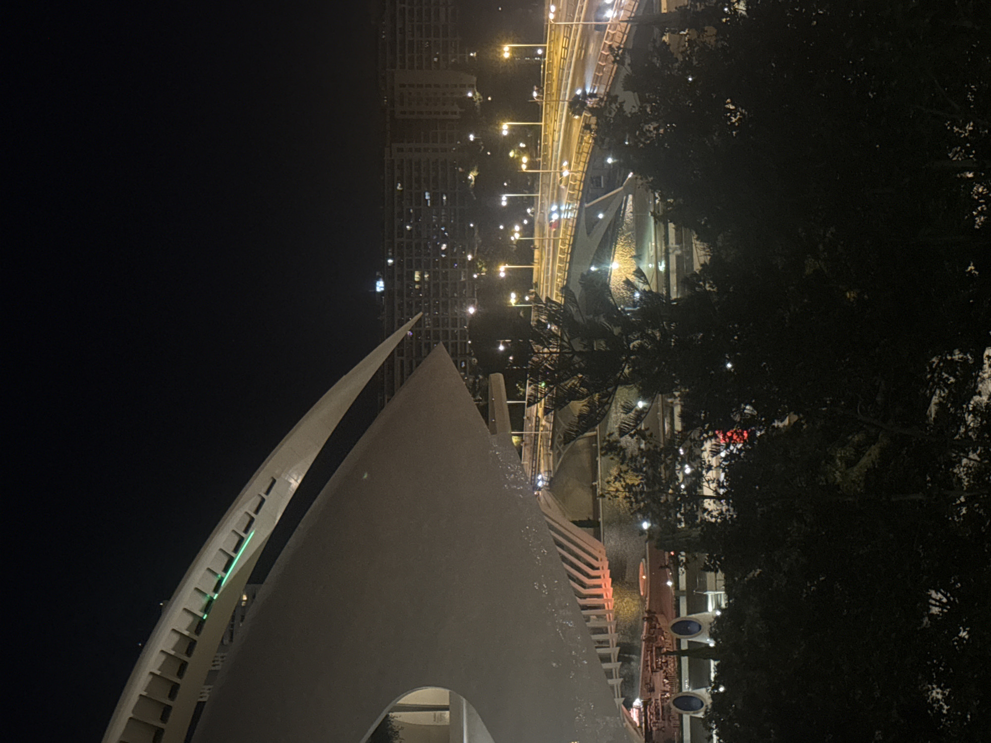 This image captures a nighttime view of the City of Arts and Sciences in Valencia, Spain. In the foreground, a large, curved white structure, part of the Hemisfèric, dominates the left side of the frame, with a section of green lighting visible along its edge. Below and to the right, a multi-level bridge is illuminated by streetlights, with cars visible on its roadway, indicating active traffic. In the middle ground, a body of water is present, reflecting some of the surrounding lights. Beyond the bridge, a cluster of trees obscures some of the view. In the background, numerous buildings are lit up, suggesting a densely populated urban area. The overall scene is one of urban architecture and infrastructure bathed in artificial light against the dark night sky.