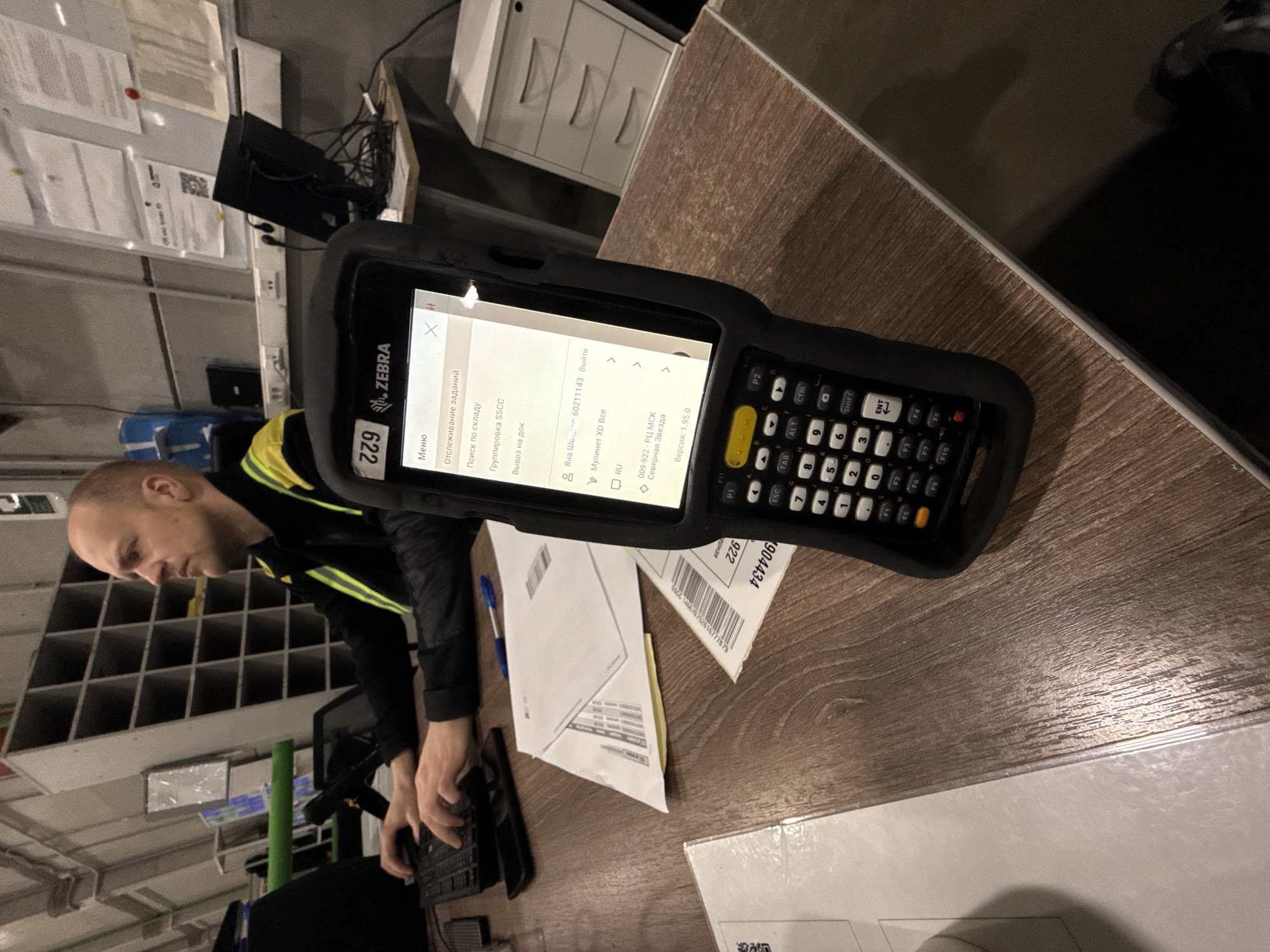 The image shows a man in Lupanovo, Russia, working at a desk. He is wearing a black jacket with yellow stripes. He is interacting with a Zebra mobile computer with a QWERTY keyboard and a screen displaying Russian text. The screen reads "Отслеживание заданий," meaning "Tracking Tasks," and includes menu options, information about "Яна Швидас," "Мулинет XD Все," and "Северная Звезда." Several documents are lying on the desk, as well as a pen and a computer keyboard. A barcode is visible on one of the documents. In the background is a wall-mounted metal rack with square compartments, office furniture, and papers pinned to a bulletin board. The desk has a wooden surface.