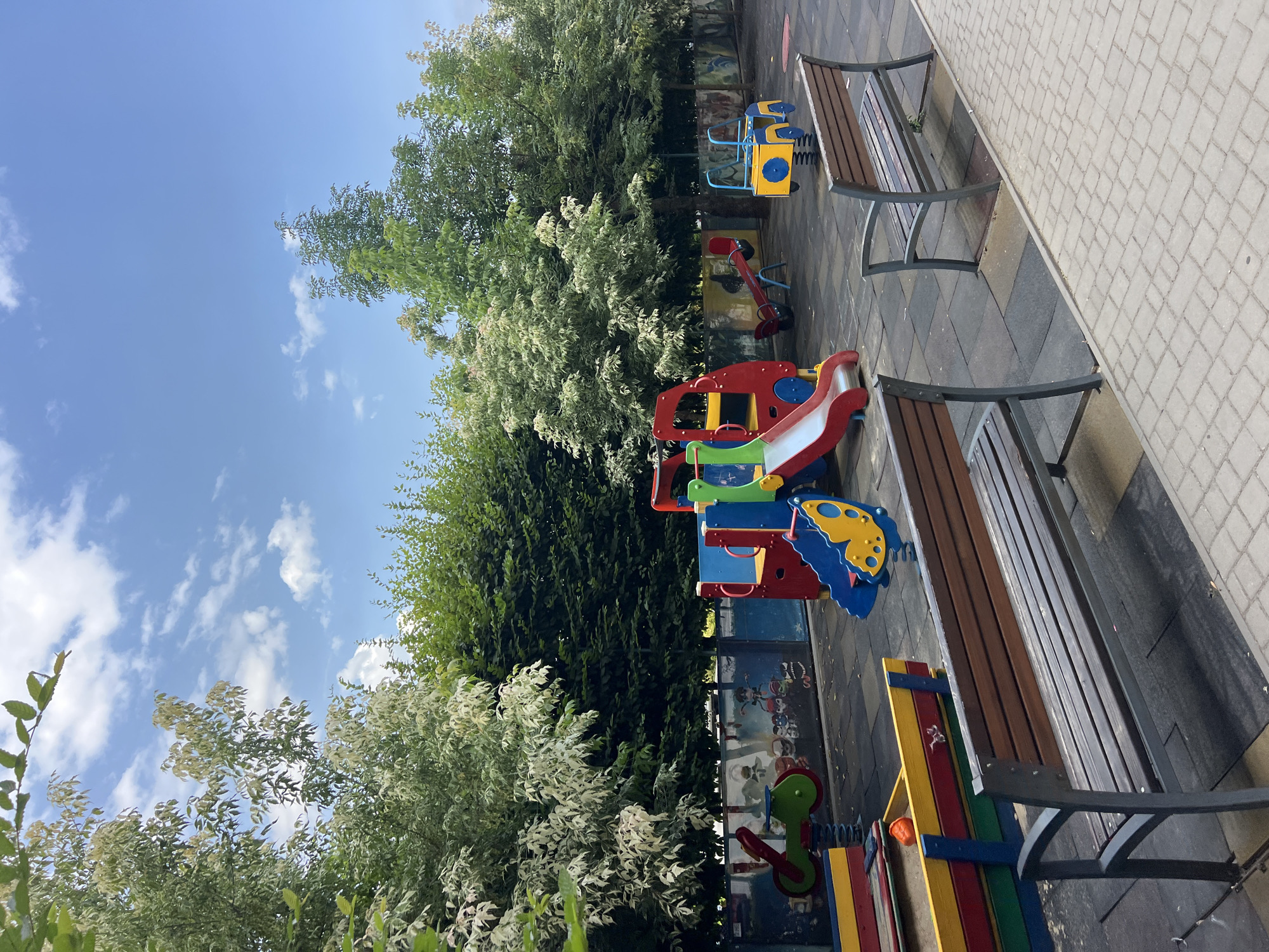 The image depicts an outdoor children's playground. In the foreground, two wooden benches with dark metal frames are positioned on a rubberized surface, adjacent to a paved walkway. Behind the benches, a colorful sandbox with red, yellow, and green trim contains sand and a single orange ball. To the left, a green and red spring rider toy is visible.
Dominating the center of the image is a vibrant, multi-colored playground structure featuring a red slide with yellow accents, blue steps, and red and yellow platforms. To the right of this structure, a red seesaw with blue seats is present, and further right, a blue and yellow spring-loaded toy resembling a small vehicle with a yellow circular detail on its side. The background is bordered by a low wall adorned with cartoon murals.
Lush green trees, some with variegated white and green leaves, line the perimeter of the playground, creating a natural backdrop against a bright blue sky dotted with scattered white clouds. The scene suggests a well-maintained public space. The location is identified as Fontanka, Ukraine. There are no people visible in the image.