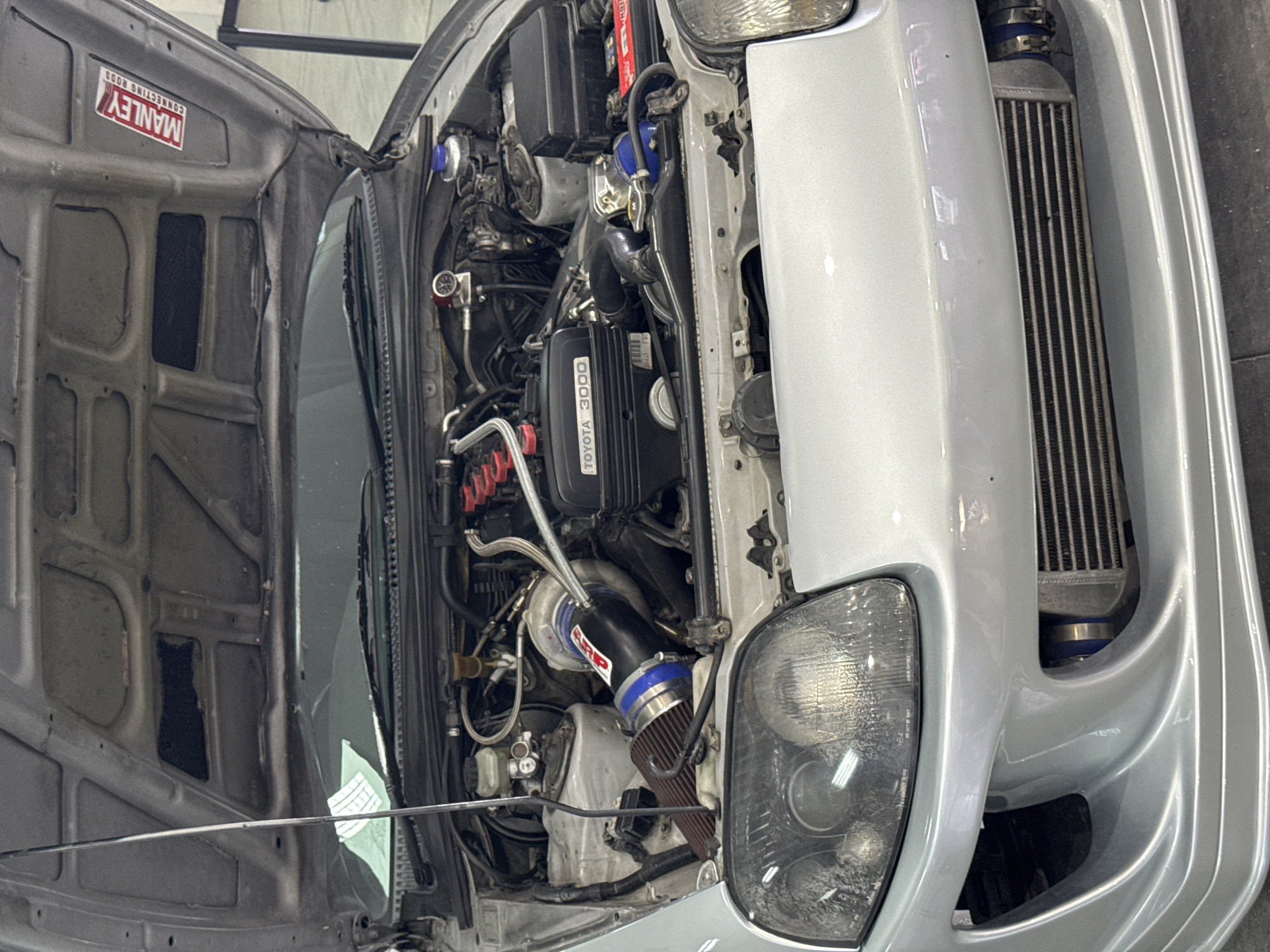 The image shows the engine bay of a silver Toyota Supra. Visible engine components include a "TOYOTA 3000" valve cover, a turbocharger with an aftermarket air intake filter, and an intercooler mounted at the front. The car has aftermarket parts like red ignition coils. The hood is open and bears a sticker for "MANLEY CONNECTING RODS." The specific location in Plovdiv, Bulgaria is not discernible from the image. There are no people visible.