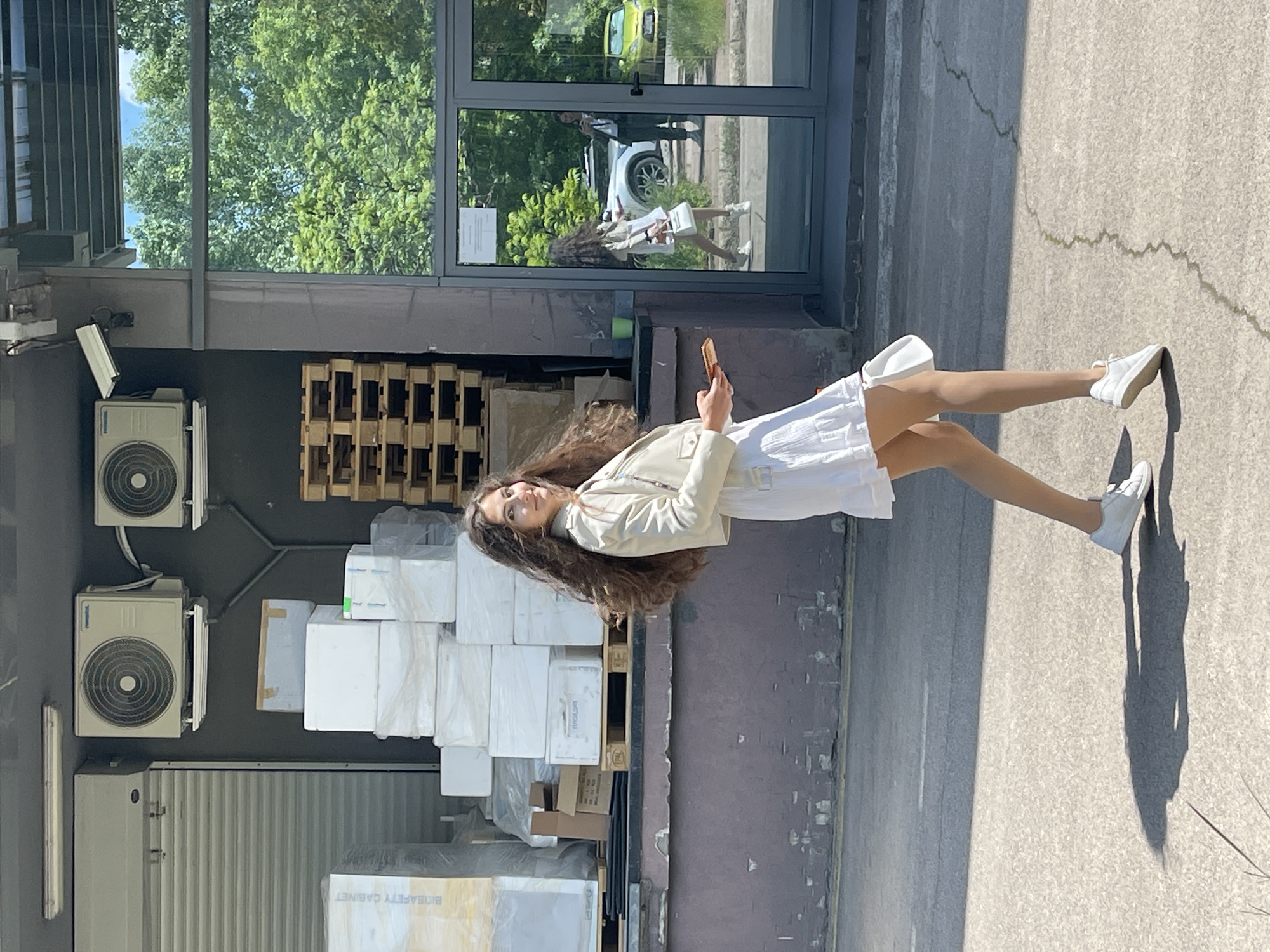 A young woman stands on a concrete sidewalk next to a building in Sofia, Bulgaria. The building is modern, with a large glass window reflecting the trees and buildings outside. The woman is wearing a white dress and light-colored jacket. She holds a smartphone in her hand. The building is near equipment, including air conditioning units and boxes on wooden pallets. The lighting indicates it is daytime. The overall scene presents an urban environment.