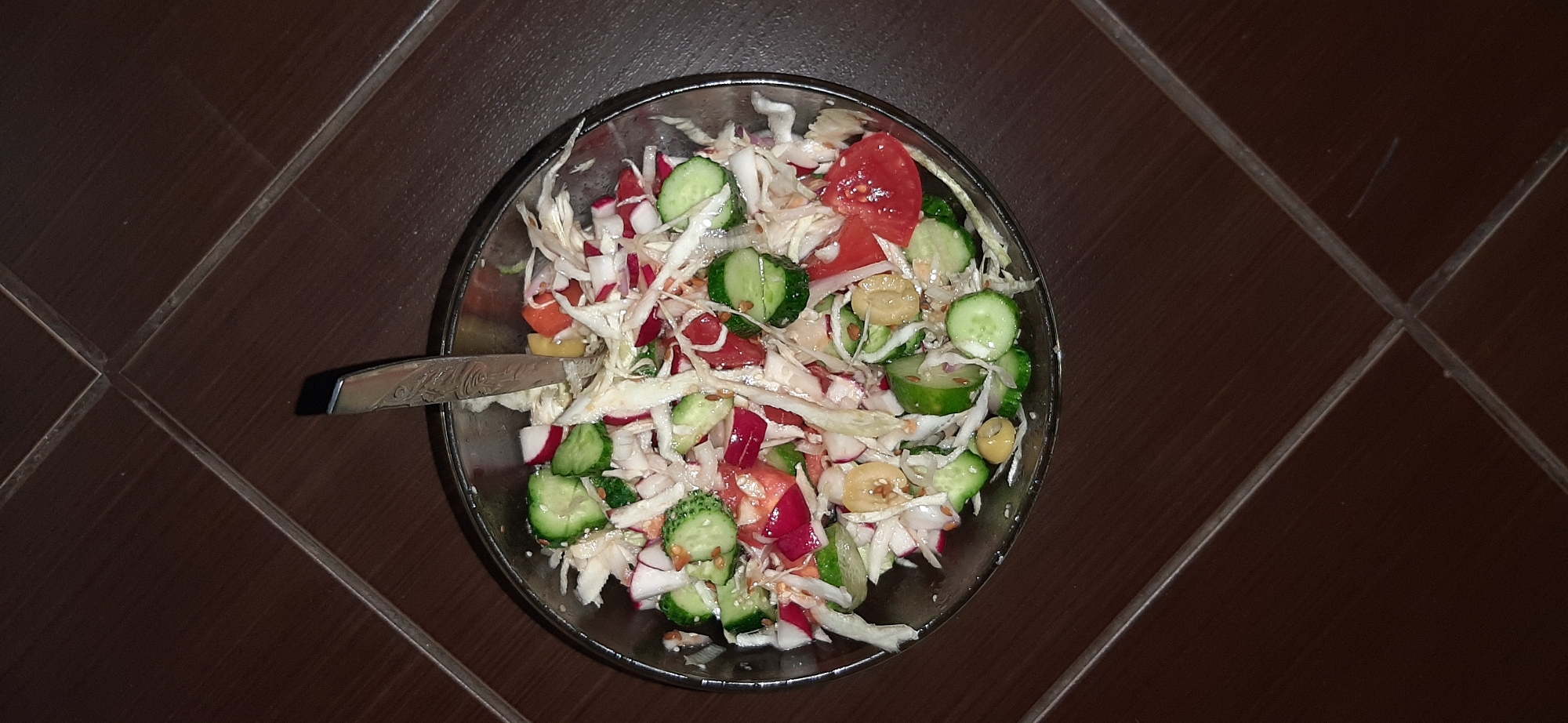 The image shows a close-up, overhead view of a salad in a clear bowl. The salad contains chopped cabbage, sliced cucumbers, diced tomatoes, sliced radishes, and what appear to be chickpeas or similar legumes. A metal fork with an engraved handle is resting in the bowl. The bowl is placed on a dark brown tiled surface, possibly a floor or countertop. The lighting suggests an indoor setting, likely at night. There is no visible indication of the location being Chernihiv, Ukraine, or any specific city.