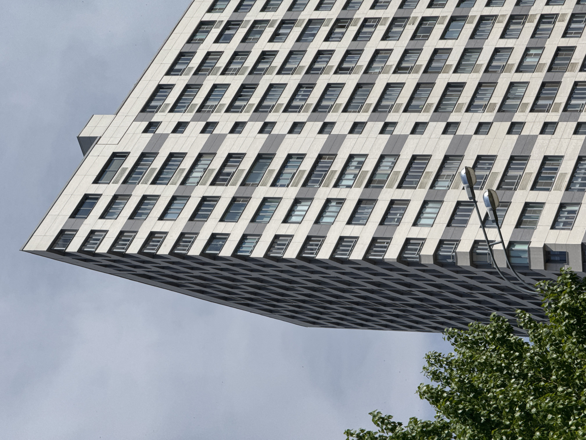 A low-angle shot shows a section of a high-rise building in Kazan, Russia, with many windows and a light gray and dark gray striped facade. The building overhangs, with the underside visible, showing a grid-like pattern of windows and dark surfaces. At the bottom right of the frame, part of a tree with green leaves is visible. A street light stands near the tree, against the background of the building. The sky in the background is overcast and gray.