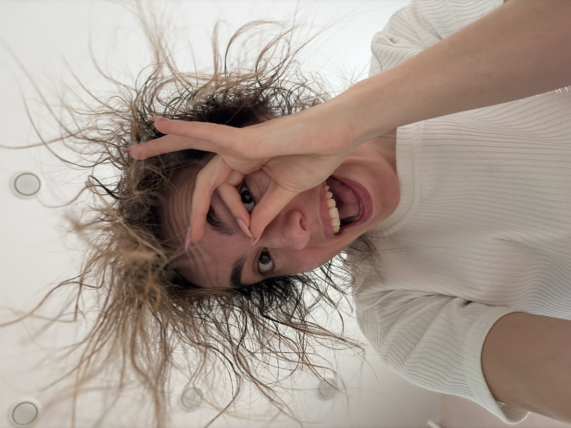The image, taken in Izhevsk, Russia, captures a dynamic, low-angle portrait of a young adult female, appearing upside down as she looks towards the camera from directly above. Her long, light brown hair is disheveled and stands wildly around her head, suggesting movement or static electricity. She is wearing a light-colored, possibly white or off-white, short-sleeved ribbed top.
Her left hand is raised to her face, with her thumb and index finger forming a triangular shape through which her right eye is visible, looking directly at the viewer. Her mouth is wide open, revealing her teeth, in an expression that conveys a sense of excitement, surprise, or playful shouting/laughing.
The background is a plain white ceiling with several circular recessed light fixtures, indicating an indoor setting. The perspective makes her hair appear to defy gravity, adding to the energetic and somewhat chaotic mood of the image. No text is visible in the frame.