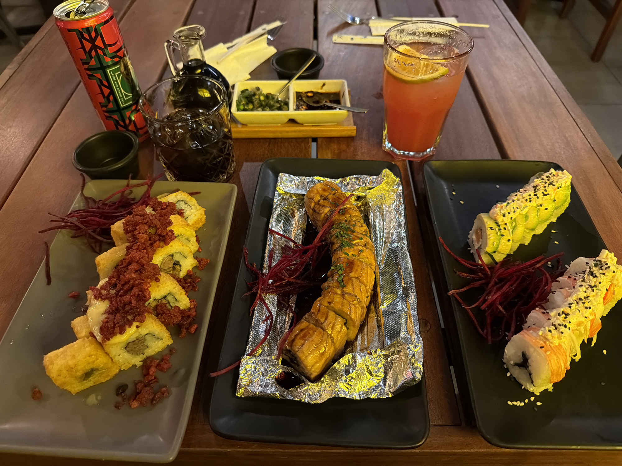 The image shows a table laden with various dishes, likely in a restaurant setting, in Unknown, Unknown. There are three plates of sushi: the left plate presents fried sushi topped with reddish-brown crumbles; the central plate holds a glazed, grilled roll on aluminum foil, garnished with red sprouts; the right plate displays two types of sushi rolls, one featuring avocado and the other containing crab stick.
Beverages on the table include a red canned drink, a glass of dark liquid, a small jar of dark sauce, and a tall glass of pink liquid with an orange slice. A small dish of green condiments and dark sauce sits near chopsticks wrapped in paper. The table is made of wood, and the background is blurry, indicating an indoor environment. No people are visible in the image.