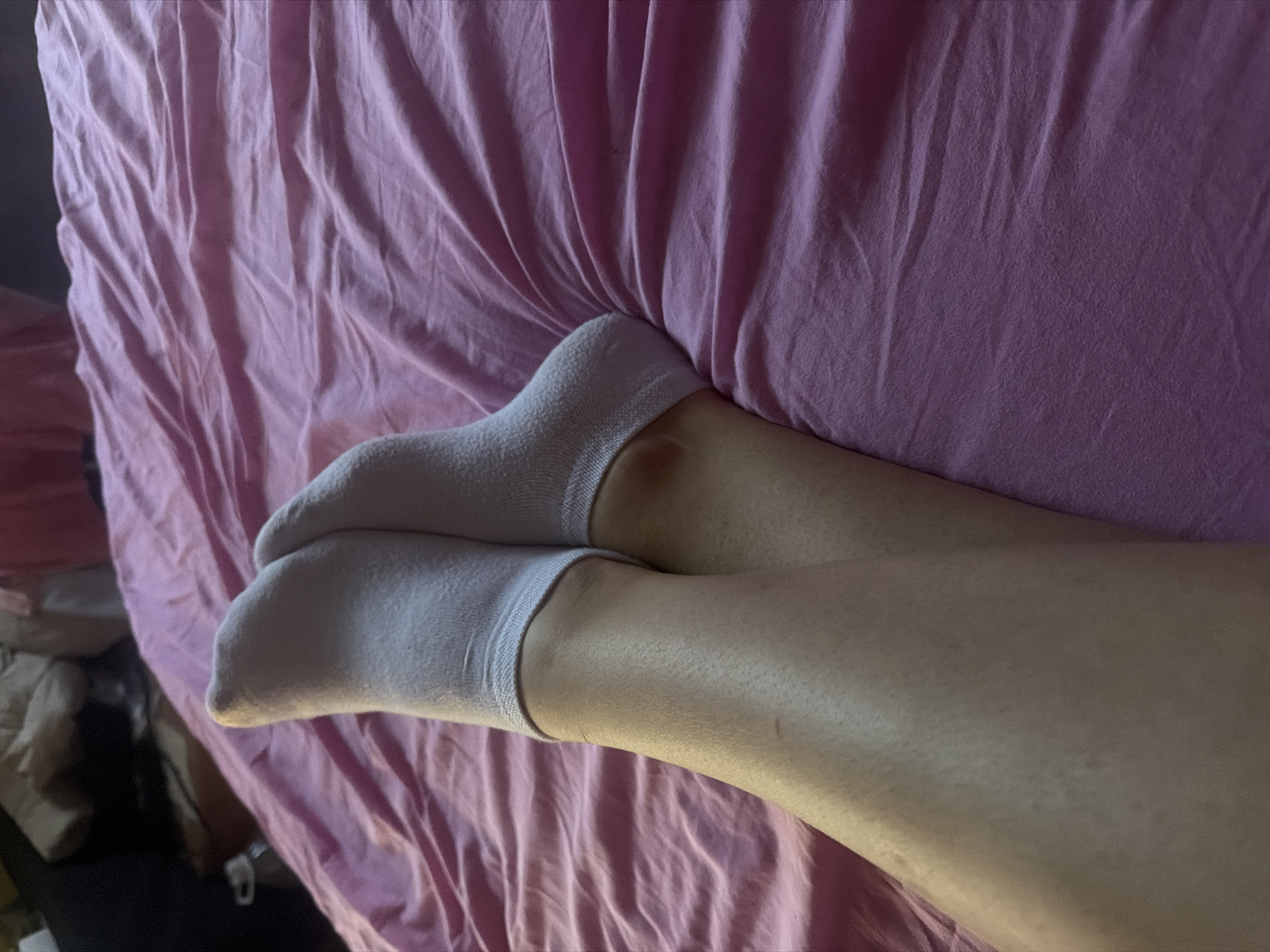 A close-up studio shot shows a pair of legs wearing white socks resting on a wrinkled pink bedsheet. The background consists of more of the pink sheet, and a glimpse of furniture and other items is visible on the left.