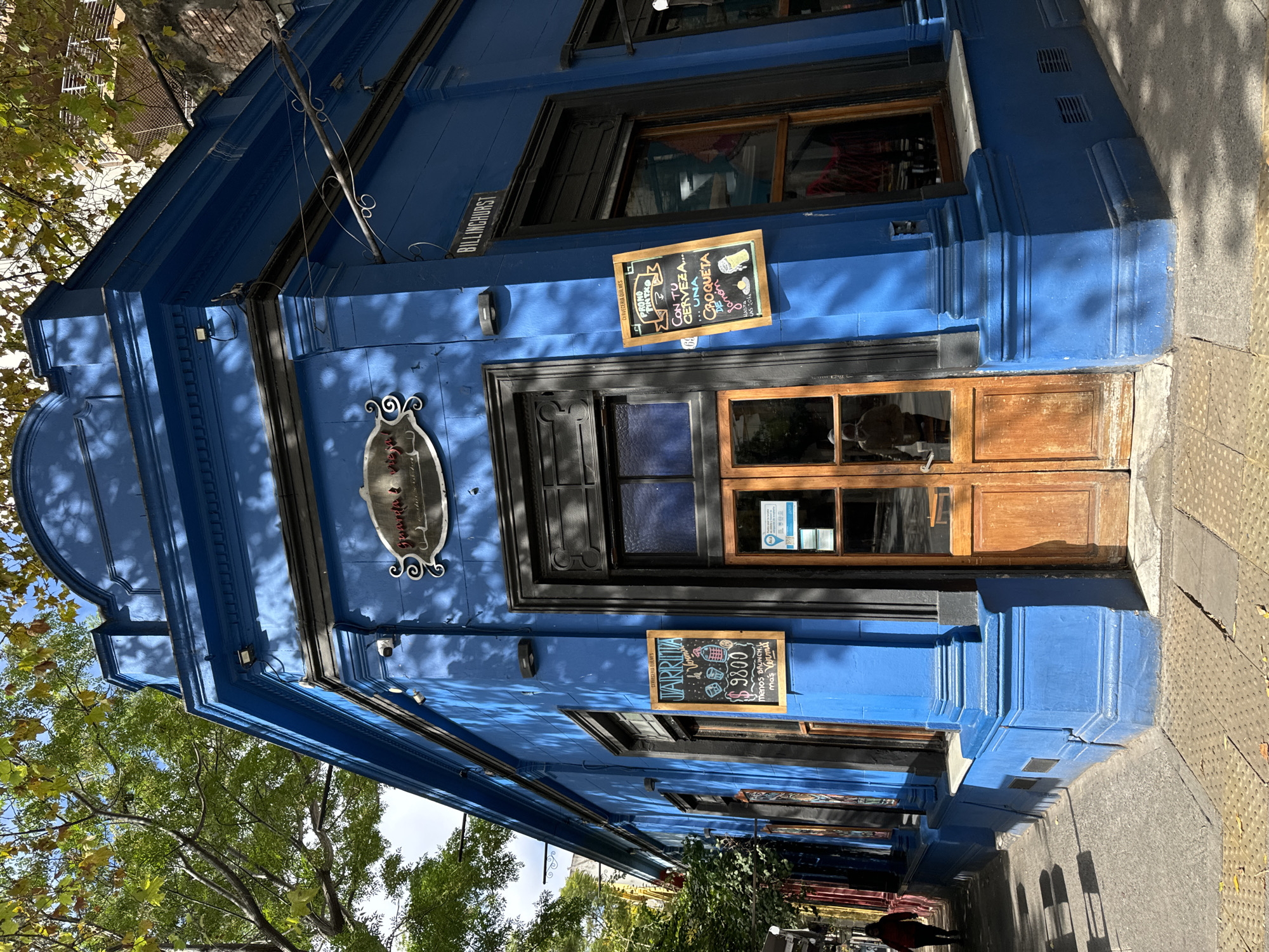 The image shows the exterior of a bright blue building, likely a business, located on Billinghurst street in Buenos Aires, Argentina. The building has dark trim around windows and doors. Signs attached to the wall advertise "JARRIVA $9800" and beer with croquettes, suggesting a bar or restaurant. One sign reads "Cerveceria Quilmes." The building has an ornamental sign with the name "Gourmets & Co." There are trees partially visible along the street. A person in a red jacket is walking on the sidewalk in the distance. The sidewalk is paved with textured concrete.