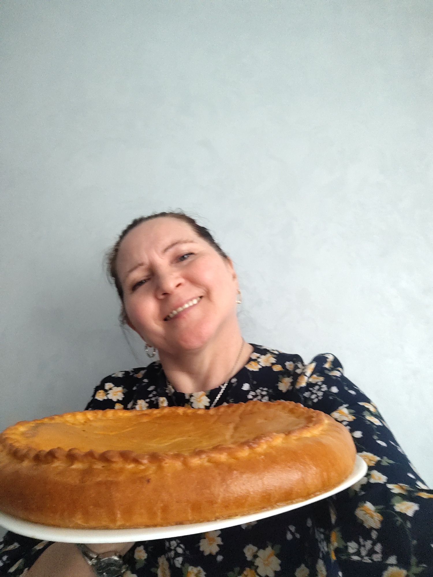 A smiling woman joyfully presents a large, homemade golden-brown pie in an indoor, close-up shot, likely taken in Sarapul, Russia. She looks directly at the camera with a wide, warm smile, revealing her teeth, and her medium brown hair is neatly pulled back. She wears a dark blue dress or top adorned with a pattern of yellow and white flowers, a silver chain necklace, and a silver watch on her left wrist. The prominent, round pie, with its distinct crimped crust, rests on a white plate, occupying the lower part of the frame. The background is a plain, light blue-gray wall. The scene captures a moment of domestic pride or hospitality, with the woman appearing delighted to share her baked creation. The lighting is bright and even, suggesting a daytime setting. No text is visible in the image.