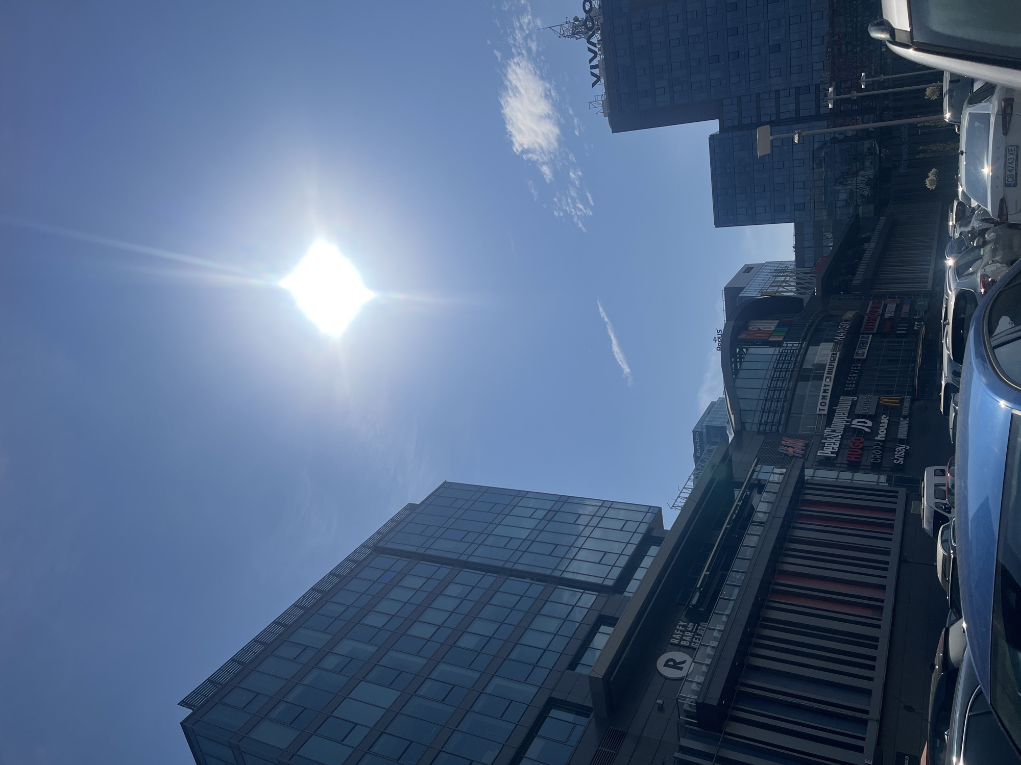 An urban outdoor scene unfolds on a bright, sunny day, featuring modern architecture and a commercial district. Several tall, glass-fronted buildings rise against a clear, vibrant blue sky dotted with a few wispy clouds. The sun, intensely bright and positioned in the upper left, creates a prominent lens flare.
The central subject is a multi-story shopping center, its facade adorned with numerous retail brand logos. Visible brands include TOMMY HILFIGER, MANGO, Peek&Cloppenburg, RESERVED, HUGO, JD, CALICE, CROPP, house, sinsay, HUMANIC, and Texanova. Other businesses identified are "RAFFY BAR AND GELATO" with an "R" logo, and a "fitness club." Further up, "VIVACOM" is visible on a separate building, and "Aurubis" can be faintly seen near the top right.
A parking area is present in the foreground and to the right, occupied by several cars. A blue vehicle is partially visible in the immediate foreground, and a white car's license plate, "CB 4743 XE," is clearly readable. The "CB" designation on the license plate confirms the location as Sofia, Bulgaria.
The scene captures a static moment in a bustling city environment, likely during late morning or early afternoon due to the high and bright sun. No specific human activity is discernible.
