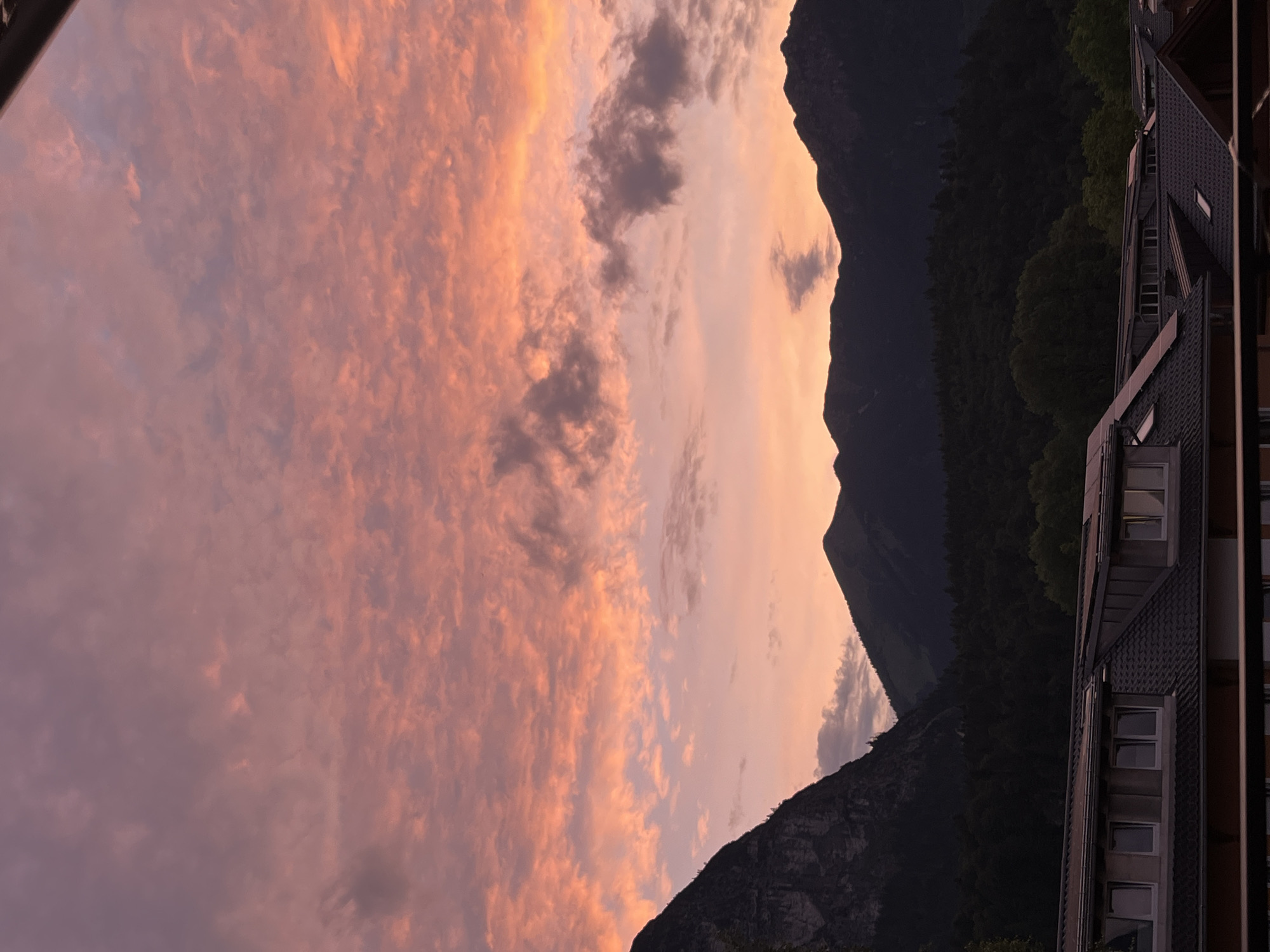 The image captures a mountain landscape at sunset. The sky is filled with clouds that are illuminated by the setting sun, displaying hues of pink and orange. In the foreground, a portion of a building with a tiled roof and several windows is visible. Behind the building, a dense forest covers the lower slopes of dark, silhouetted mountains. The scene appears to be a view from a higher vantage point, looking out over a residential area towards the mountains and sky. The location is identified as Garmisch-Partenkirchen, Germany, a town known for its alpine scenery.