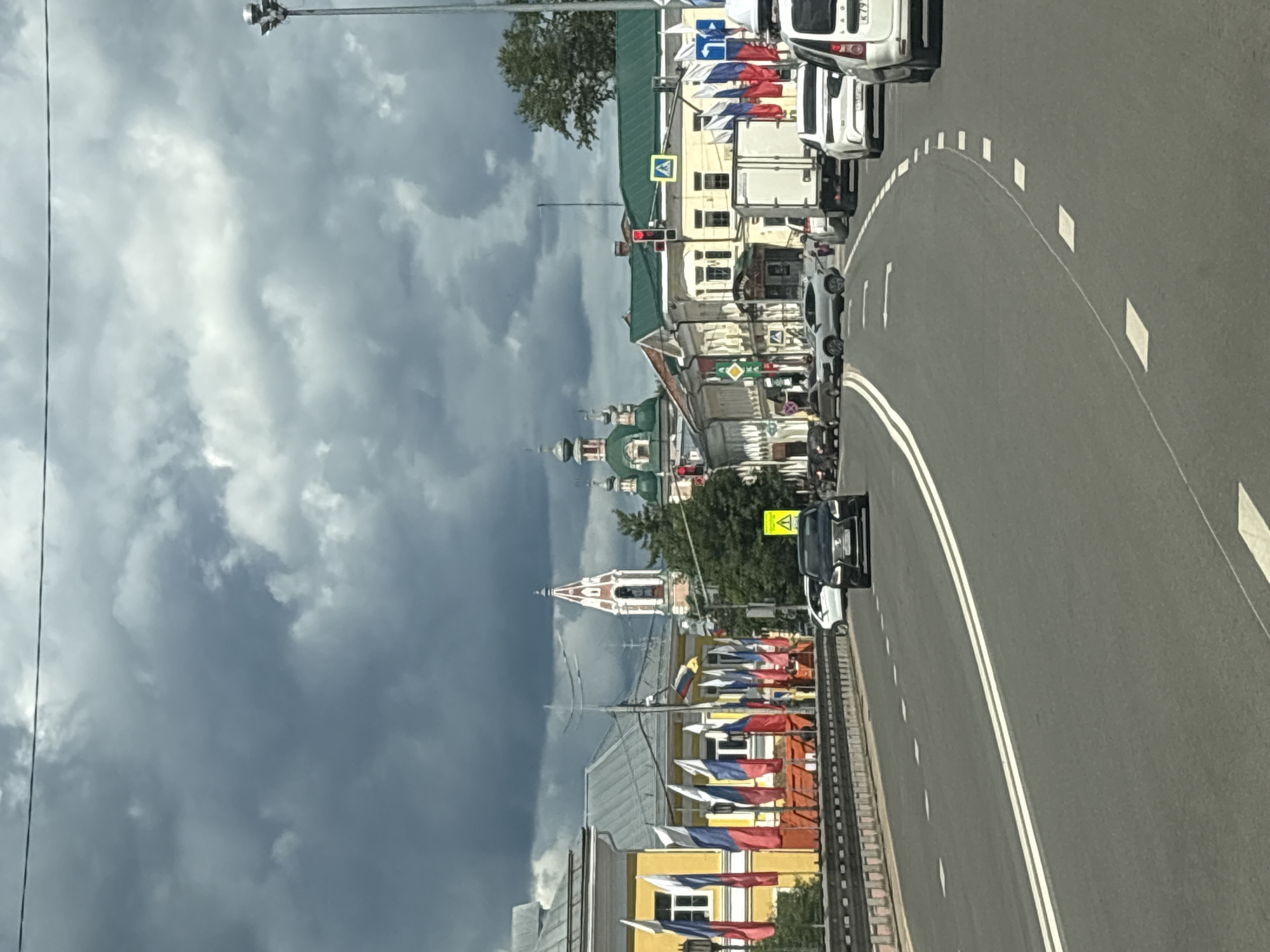 The image shows a street scene in Pereslavl-Zalessky, Russia, on a cloudy day. The view is from a vehicle traveling on a road with double white lines. On the left side of the road, there is a yellow building decorated with numerous Russian flags on flagpoles. In the background, several church domes and spires are visible, characteristic of Russian Orthodox churches, including a prominent bell tower with a white and red design. On the right side of the road, there are other buildings, traffic lights, and several vehicles, including a white truck and a white SUV. The sky is filled with dark, dramatic clouds.