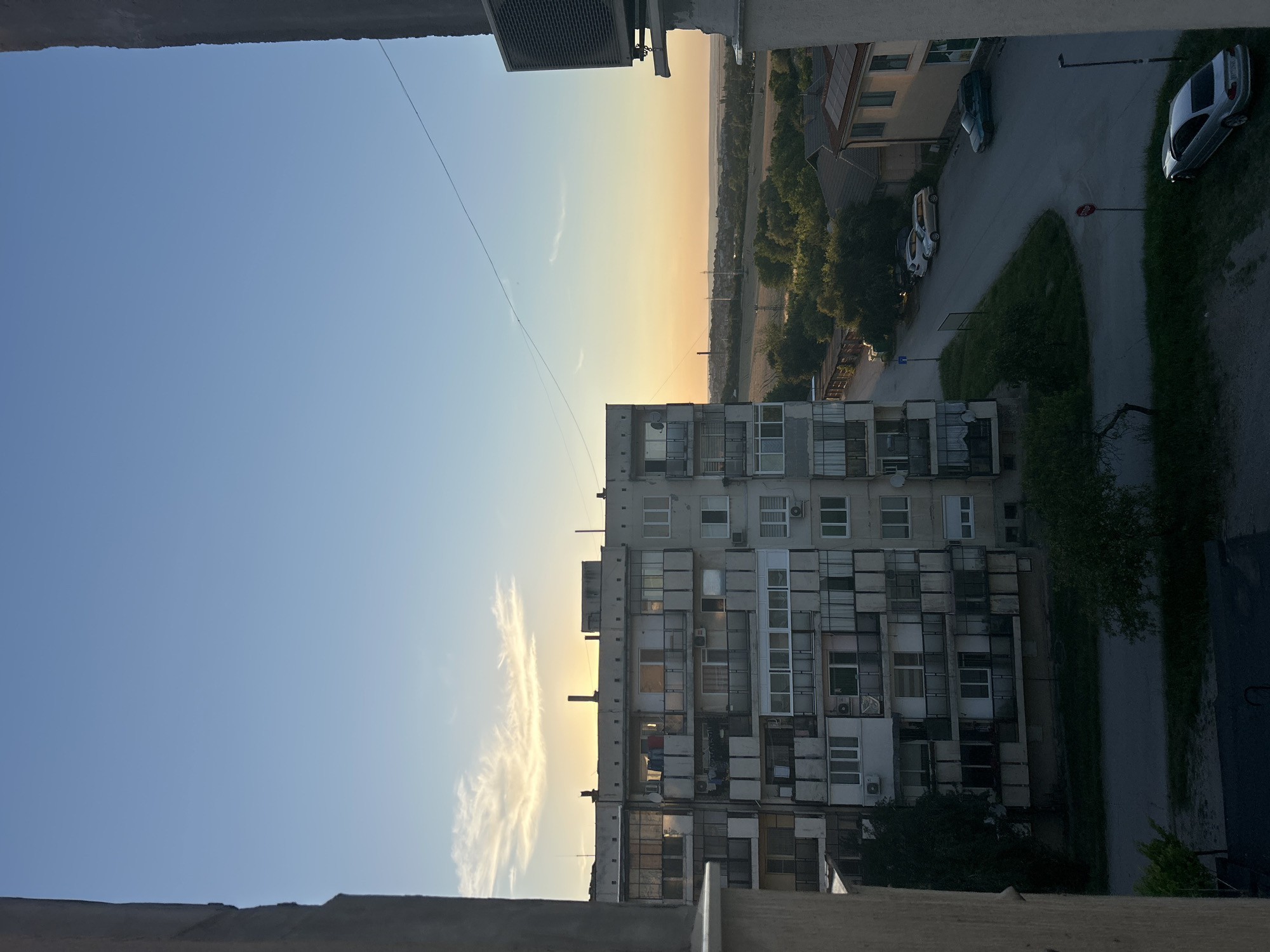 An exterior, eye-level, medium shot captures a view from a window of a multi-story apartment building at dusk in an urban setting. The sky above is a gradient of light blue to soft orange as the sun sets, with a few wispy clouds. To the left, a portion of the apartment building is visible, featuring numerous balconies with laundry hanging out to dry and satellite dishes. The building is gray and has a worn, utilitarian appearance. In the foreground, the concrete frame of the window is visible, creating a border around the scene.
To the right of the apartment building, a street curves through the neighborhood. Several cars are parked along the street, including a silver sedan in the foreground, a green hatchback, and two lighter-colored sedans further up the road. Houses with tiled roofs are visible beyond the street, surrounded by trees and vegetation. In the distance, a flat, treed landscape stretches to the horizon, with some distant structures and power lines. The overall atmosphere is quiet and serene, characteristic of the end of a day.