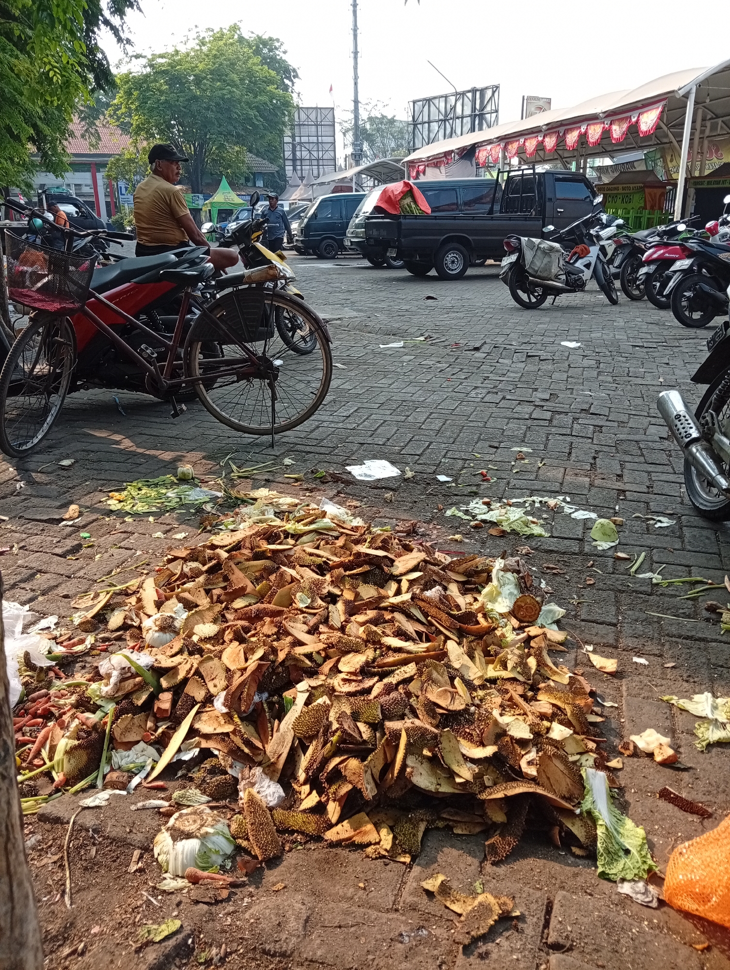 The image depicts an outdoor scene in the city of Larangan, Indonesia, likely a market area. In the foreground, a large pile of discarded food waste, primarily durian rinds and vegetable scraps, is visible on a cobblestone pavement. A red bicycle with a basket is parked to the left, partially obscuring a motorcycle. Further back, several motorcycles and a black pickup truck are parked. A man wearing a yellow shirt and a black cap is seated on a motorcycle. In the background, a market stall with a white roof and red and white banners is visible, along with other vehicles and buildings. The lighting suggests it is daytime, with the sun casting shadows. The overall scene conveys a sense of a busy commercial area where waste disposal is a concern.