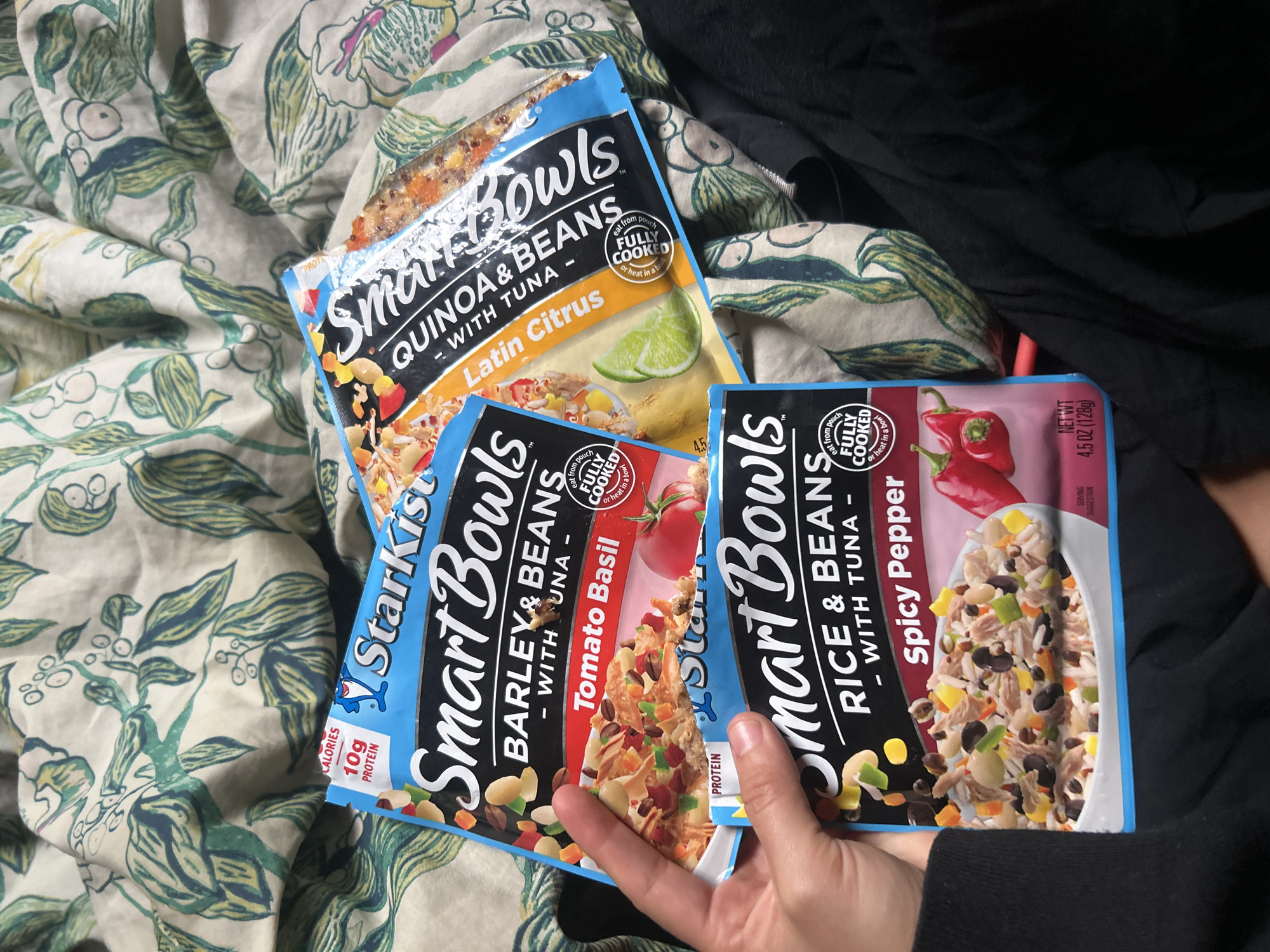 Here's a description of the image:
The image shows three pouches of StarKist Smart Bowls arranged on what appears to be a bed with a floral patterned sheet. A person's hand is visible, holding the pouch of "Rice & Beans - with Tuna - Spicy Pepper" and partially covering the "Barley & Beans - with Tuna - Tomato Basil" pouch. The third pouch, "Quinoa & Beans - with Tuna - Latin Citrus," sits on top. The pouches are all pre-cooked and ready to eat. Part of a dark garment is visible in the top right corner. The setting appears to be indoors. The location is not indicated in the image.