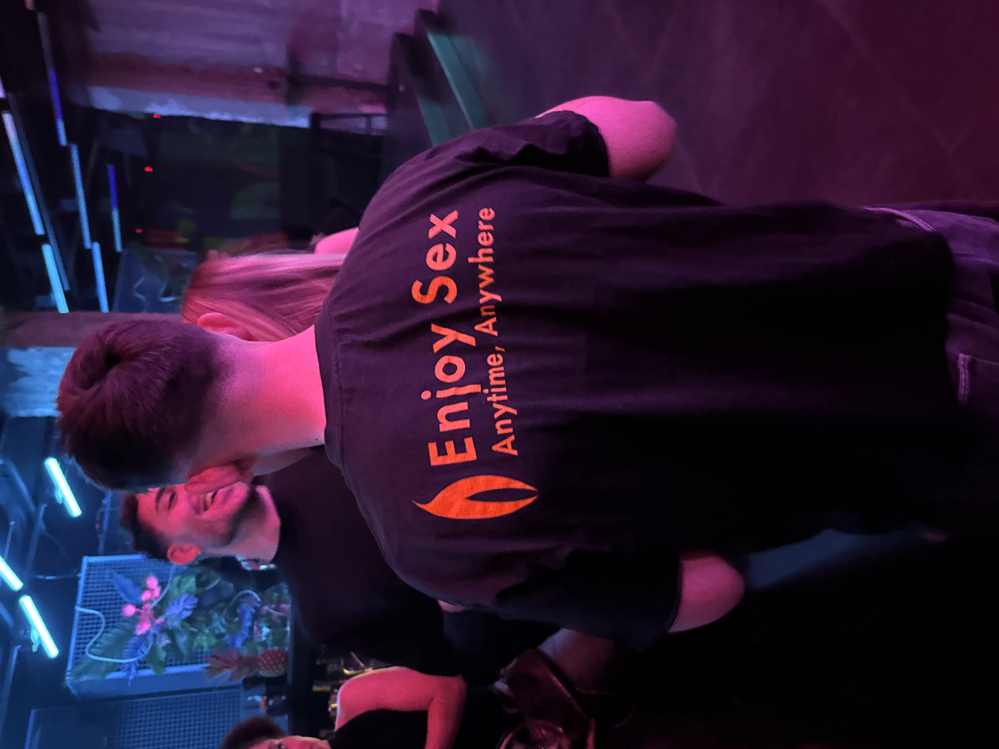 Here is a description of the image:
A medium shot captures a man wearing a black t-shirt with the words "Enjoy Sex Anytime, Anywhere" printed in orange. The t-shirt also features a leaf-like design. The man has short, dark hair and is standing with his back to the camera. Beside him is a woman with blonde hair. In the background, another man wearing a black shirt can be seen. The location appears to be a bar or nightclub setting, with blue lights and a decorative floral display on the left. The lighting is dim and colorful, with a mix of blue and purple hues.