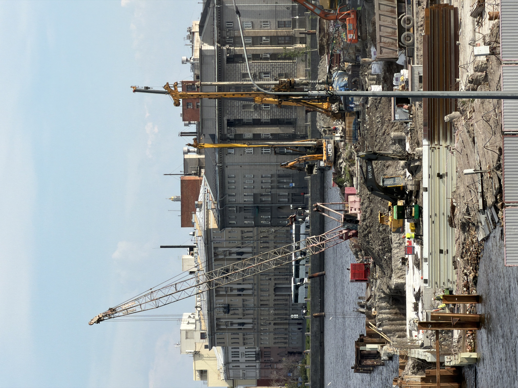 This image presents an active construction site situated on the bank of a wide urban waterway, consistent with the Neva River or a major canal in Saint Petersburg, Russia.
In the immediate foreground, a metal corrugated fence partially screens a disturbed ground area characterized by exposed earth, rubble, and various construction materials. The midground is dominated by heavy machinery and ongoing work. A large, red-and-white lattice boom crane with its boom inclined upwards is positioned on the left side of the site. To its right, two tall, yellow piling rigs are prominent, one clearly labeled "JCB," indicating foundation work. Multiple excavators, including a black-and-yellow "Volvo" machine, are present on the site, some with their buckets positioned for excavation or material movement. Piles of excavated soil, stacks of steel plates, precast concrete elements, and various pipes are scattered across the ground. Sheet piling extends into the water on the left, forming a retaining structure. Several construction workers, identifiable by high-visibility vests and hard hats, are visible on the ground, performing tasks. A large, tan-colored dump truck, loaded with earthen material, is parked on the right side of the site.
Across the waterway, a stone embankment with streetlights lines the bank. Behind this, a series of multi-story historical buildings in classical architectural styles form the urban backdrop. These structures feature columns, regularly spaced windows, and various facades, ranging from light cream to darker grey. Several white buses are parked along the embankment. The sky above is light blue with scattered white clouds, indicating clear daytime conditions. The overall scene depicts a significant urban development or infrastructure project.