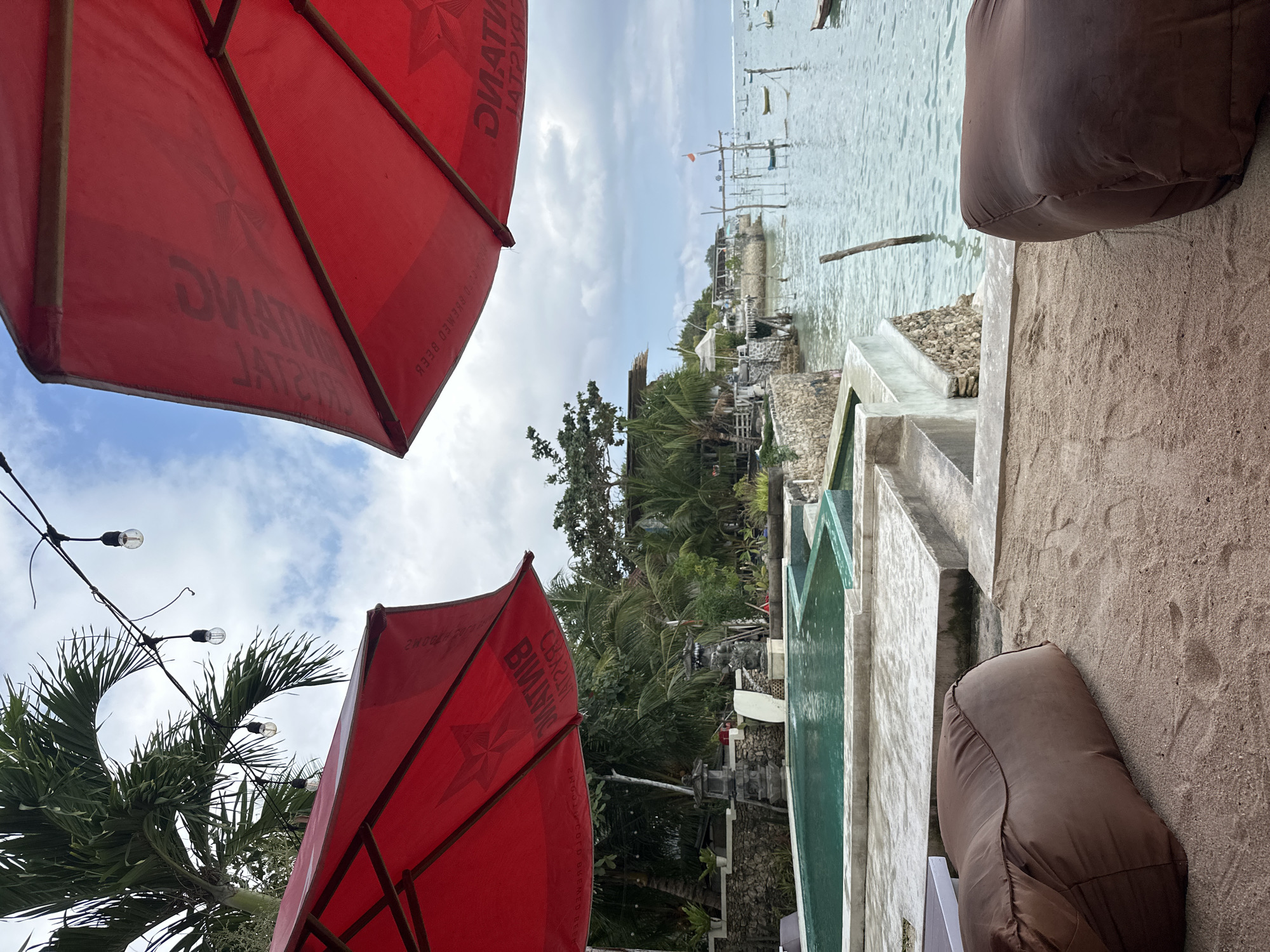 This image depicts a serene beachfront resort area in Nusa Penida, Indonesia. Two large red umbrellas dominate the foreground, casting shadows over a sandy area. In the middle ground, a turquoise swimming pool is visible, bordered by concrete structures and lush tropical vegetation. Beyond the pool, the ocean stretches out, dotted with small boats. The sky above is a mix of blue and white clouds. The scene conveys a relaxed and tropical atmosphere, typical of a vacation destination.