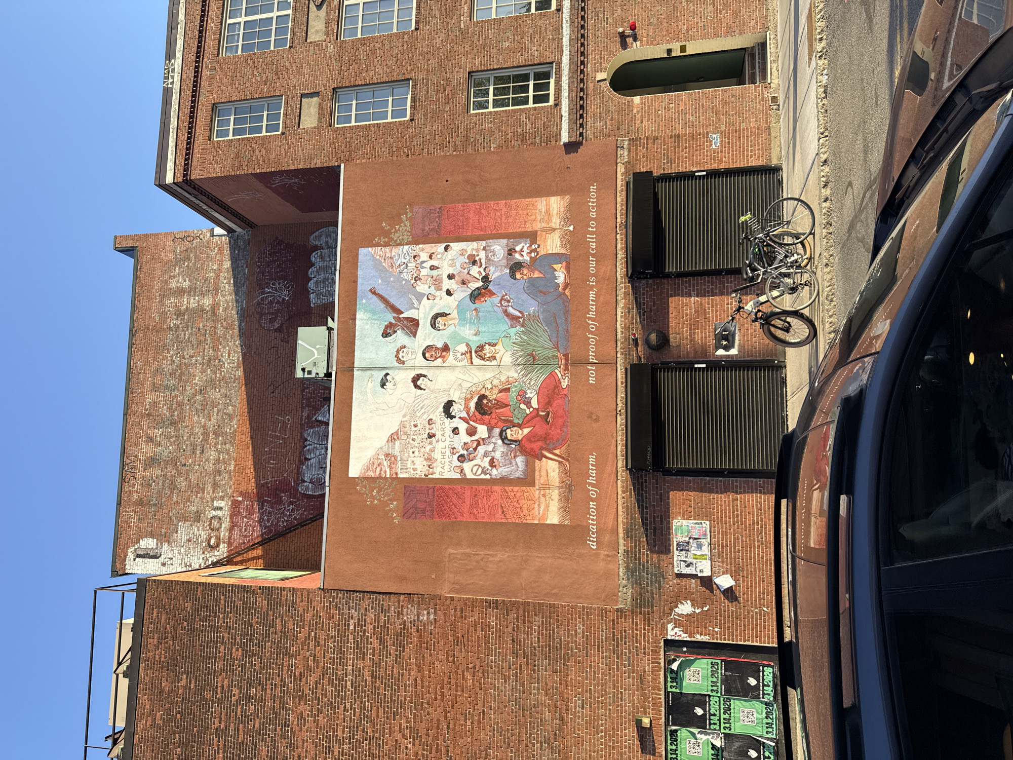 This image shows a mural on a brick building in Cambridge, Massachusetts, USA. The mural has several panels, with the largest panel showing a colorful illustration of people of various ethnicities. Some figures are depicted with tools and plants, suggesting agricultural or environmental themes. The words "Rachel Carson WAS RICH" are visible on one of the mural panels. Written below the mural are the words "dication of harm," and to the right, "not proof of harm, is our call to action." The mural is painted on the side of a building with brick construction and windows. There are bike racks with bicycles parked against the wall adjacent to the mural. The lower part of the building displays posters and graffiti. A car is visible on the right side of the image, suggesting this is an outdoor urban setting. The mural appears to be located in an urban environment, with brick buildings and visible infrastructure.