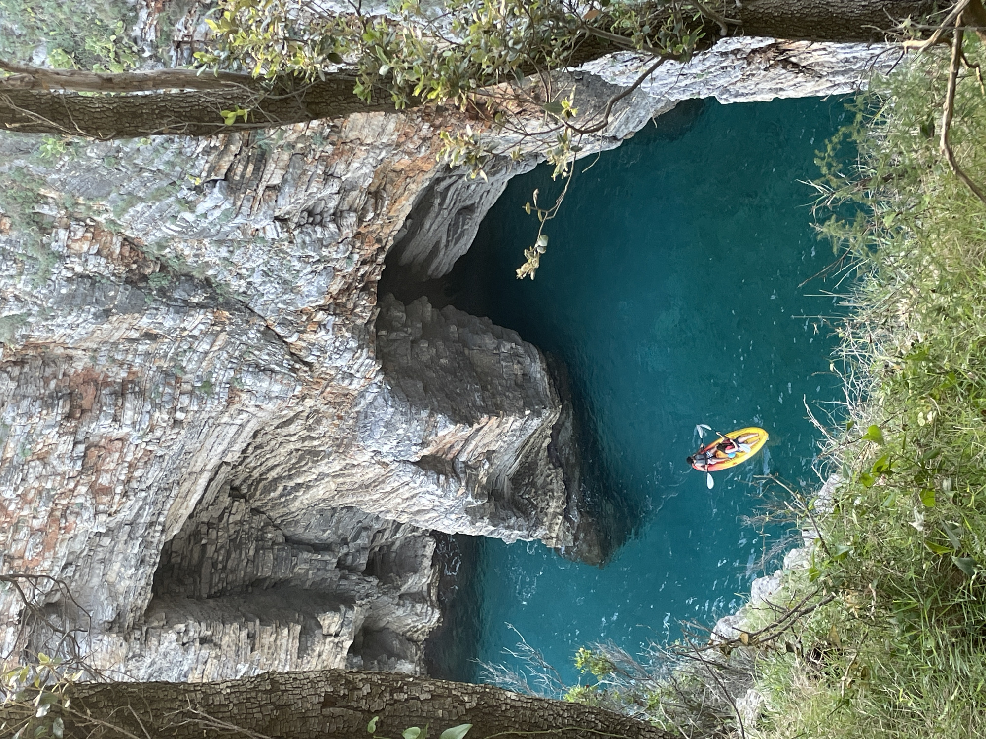 An eye-level, medium-long shot, oriented counter-clockwise, captures a narrow, turquoise cove in Budva, Montenegro. Two people are in a yellow kayak. Gray, sedimentary rock forms a cliffside to the left. Green vegetation covers the right side of the cove. The kayak floats in the center. Vegetation hangs at the top. A portion of a tree is at the bottom.