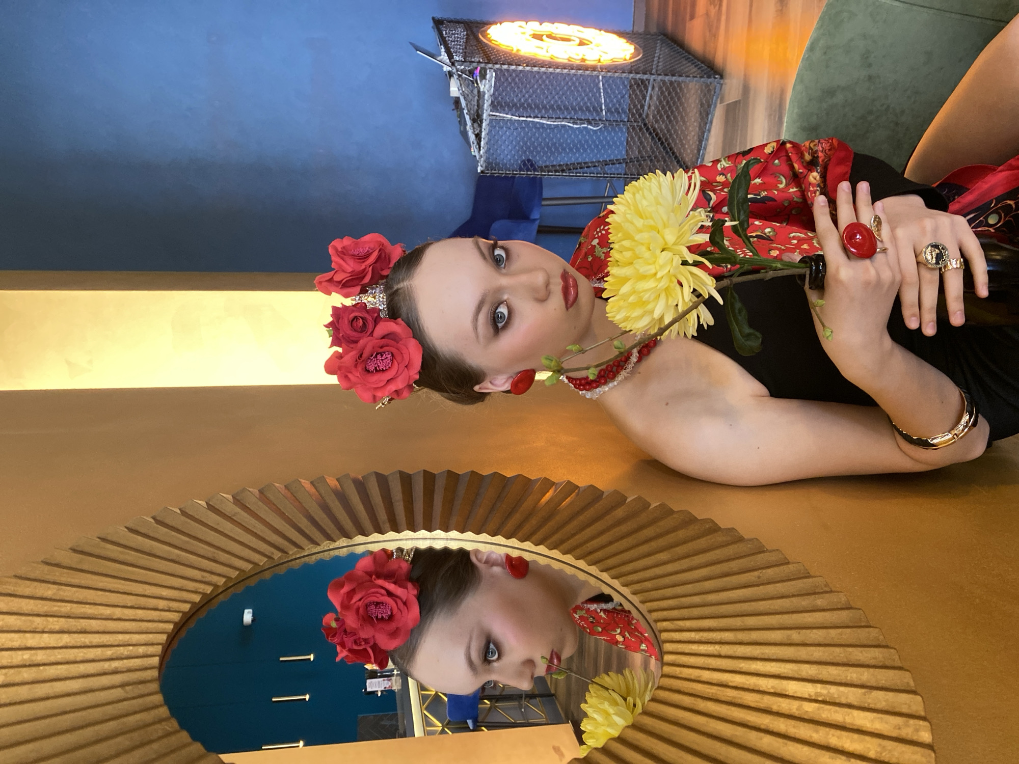A woman in elaborate attire lies on a surface next to a round, gold-rimmed mirror in what appears to be an interior setting, potentially a studio or designed space. The woman is wearing a black dress, a red shawl patterned with what looks like celestial or floral designs, a red beaded necklace, a gold bangle, and multiple statement rings. Her hair is adorned with a crown of red roses. In her hands, she holds a single yellow chrysanthemum. Her reflection is visible in the mirror, revealing more details of the room, including a blue wall and a decorative light fixture in a metal cage-like structure. The location of the photo, as stated, is Yekaterinburg, Russia.