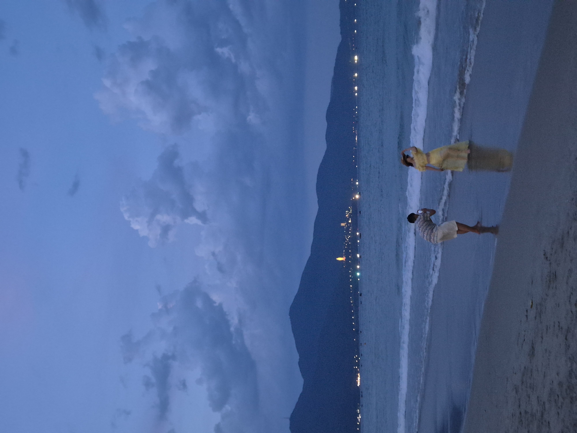 At dusk on a sandy beach in Đà Nẵng, Vietnam, a man in a striped shirt and shorts stands in the shallow water, holding a camera to his face, seemingly photographing a woman. The woman, wearing a yellow dress and a hat, stands further into the water, her hand shielding her eyes as she looks out toward the ocean. Gentle waves lap at their feet. In the background, a line of mountains is silhouetted against a twilight sky filled with dramatic, dark clouds. The distant city lights twinkle along the coastline, and a lone illuminated structure, possibly a pagoda, stands prominently on the hillside. The sea is a deep, muted blue, reflecting the dim light.