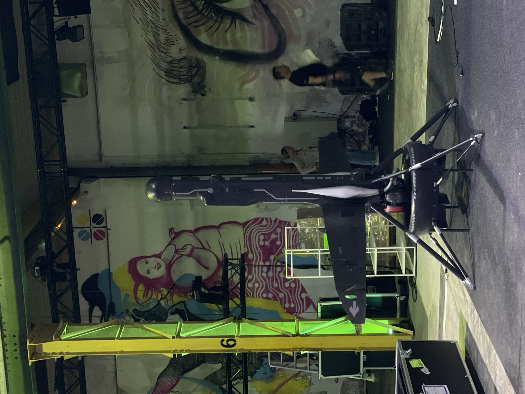 The image shows an indoor setting with a painted wall featuring a mural of a woman, along with abstract designs and color blocks, as well as what seems to be graffiti above. In front of the wall is a piece of machinery that might be a drone or a mock-up of a futuristic aircraft. It's black and streamlined, sitting on a platform or stage. A yellow structural beam is visible, possibly part of the building's architecture, and some equipment cases sit on the floor. In the background, indistinct human figures can be seen sitting at what appear to be desks or work stations.