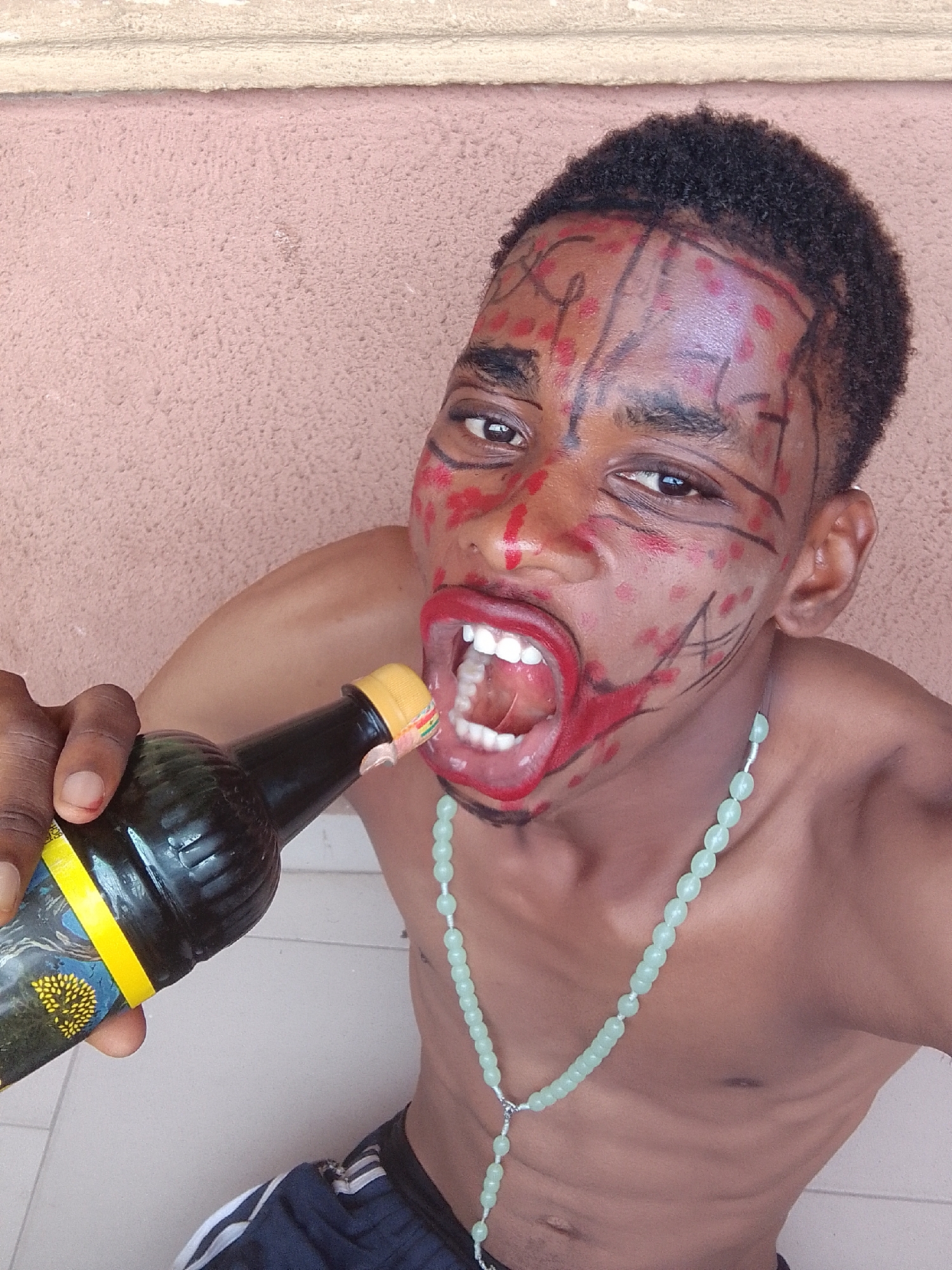 A young person, appearing to be male, with dark skin and short, dark, curly hair, is depicted in a close-up, slightly angled shot. Their face is elaborately painted with red dots and black lines/scribbles on the forehead, cheeks, and around the eyes, creating a tribal or artistic motif. Their lips are painted a vibrant red and exaggeratedly open, showcasing healthy white teeth and a visible tongue. The overall facial expression is intense and directed at the viewer.
The person is shirtless, revealing a lean upper torso and neck. A long, light green beaded necklace, resembling a rosary or mala, is draped around their neck and hangs down over their bare chest.
In their left hand, they hold a dark-colored bottle with a yellow cap. The bottle features a prominent yellow label that includes a design of a stylized yellow tree or plant against a dark blue background. The bottle is positioned directly in front of their widely open, painted mouth, suggesting either the act of drinking or a dramatic, posed gesture.
The background consists of a textured, reddish-brown wall, topped by a lighter, off-white concrete ledge or trim. Light-colored, smooth floor tiles are visible at the bottom edges of the frame. The lighting appears bright and even, but no specific time of day or outdoor weather conditions can be determined. There is no clearly legible text on the bottle or in the background. The overall impression is one of a theatrical performance or artistic expression.