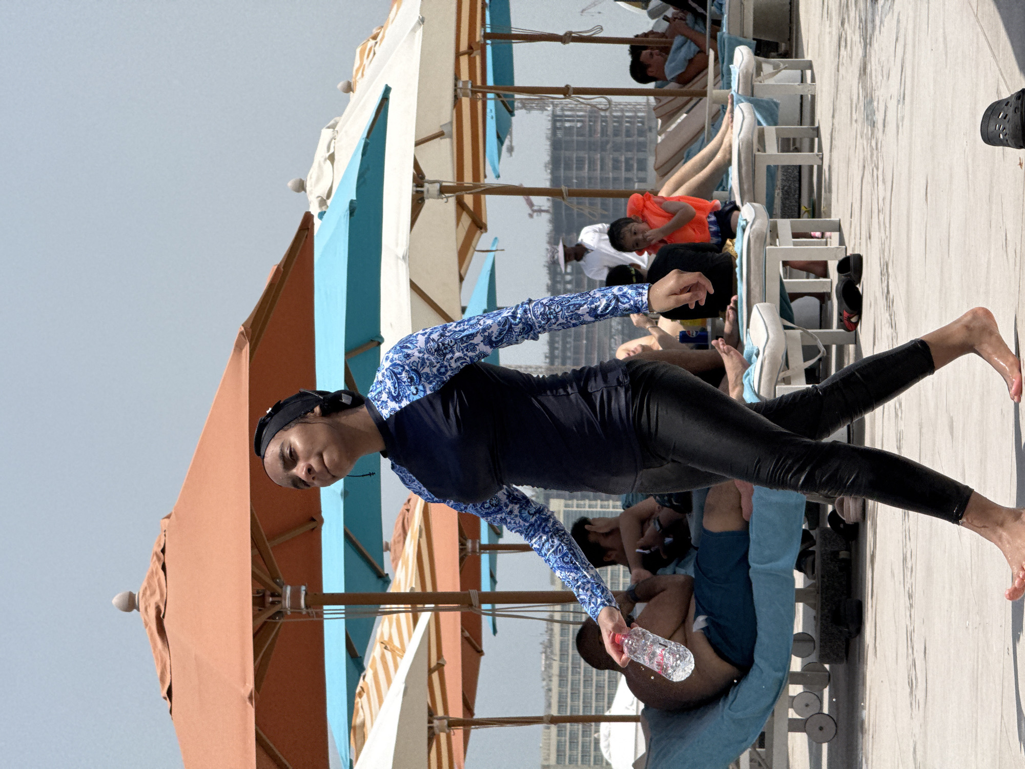 In an outdoor scene at a resort in the United Arab Emirates, a young woman in a blue and white patterned rash guard, black cap, and black leggings walks across a wet tiled surface. She is holding a clear plastic bottle of water. In the background, several people are relaxing on lounge chairs under large beach umbrellas of various colors, including orange, blue, and white. Some of the lounge chairs are occupied by individuals wearing swim trunks or bathing suits. Construction is visible in the distance, with cranes and buildings under development. A child in an orange life vest is also present, interacting with an adult. The overall environment suggests a warm, sunny day at a beach or poolside resort.
