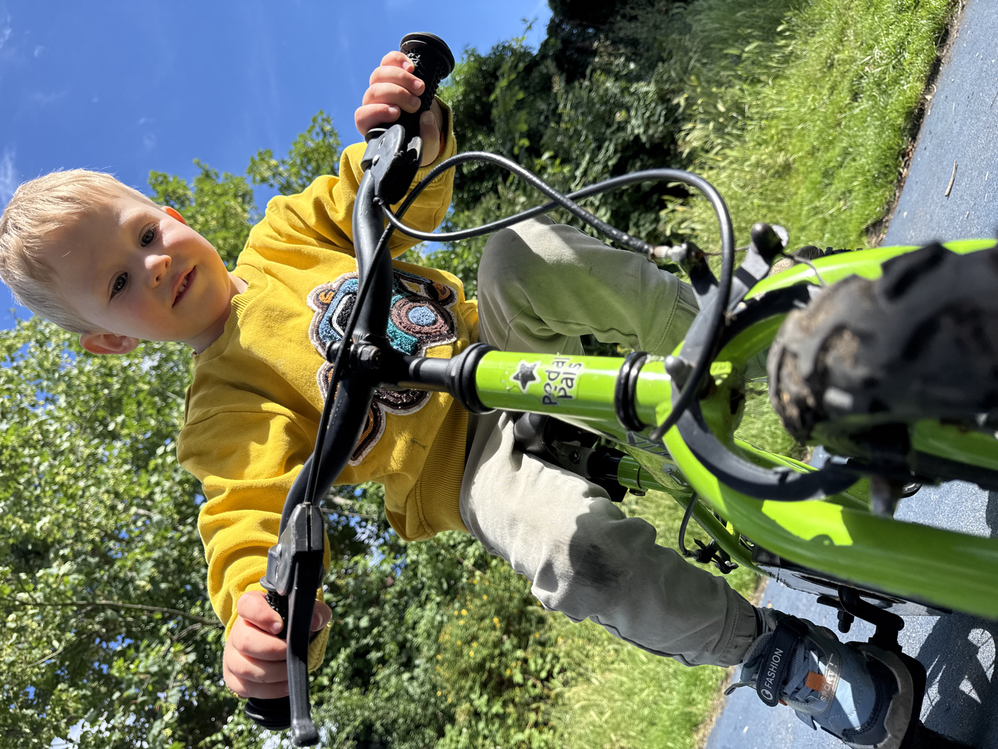A young blond boy with blue eyes is riding a bright green bicycle. He is wearing a yellow sweatshirt with a patterned design and light-colored pants. His hands are gripping the handlebars, and his feet are in sneakers. The bike has black grips and what appear to be brake levers. On one of the handlebars, there is visible text that reads "Tommi Tippi".
The boy is looking towards the camera with a slight smile, suggesting he is enjoying himself. The scene is outdoors, with lush green trees and grass in the background. The sky is a clear, bright blue, indicating it is daytime and likely sunny. The perspective of the shot is from the boy's point of view, looking down at him and the handlebars of his bike. The overall impression is a cheerful and active moment of a child learning to ride or enjoying a bike ride on a pleasant day.