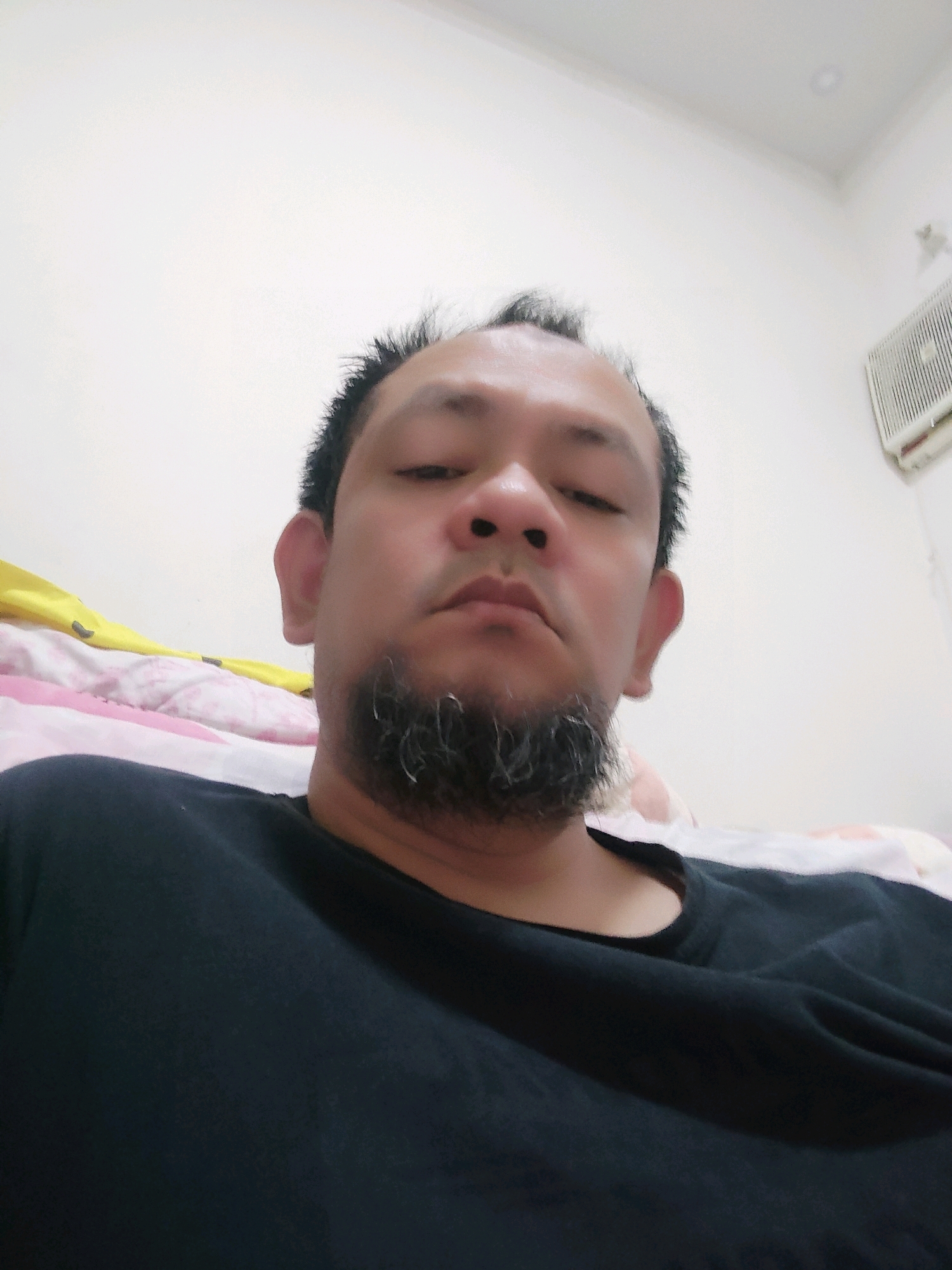 A man with thinning hair and a goatee is pictured in a selfie taken indoors. He is wearing a black t-shirt and appears to be lying down, with a pillow and blanket visible behind him. The background shows a plain wall and an air conditioning unit. The image does not contain enough information to determine the exact location within Bogor, Indonesia.