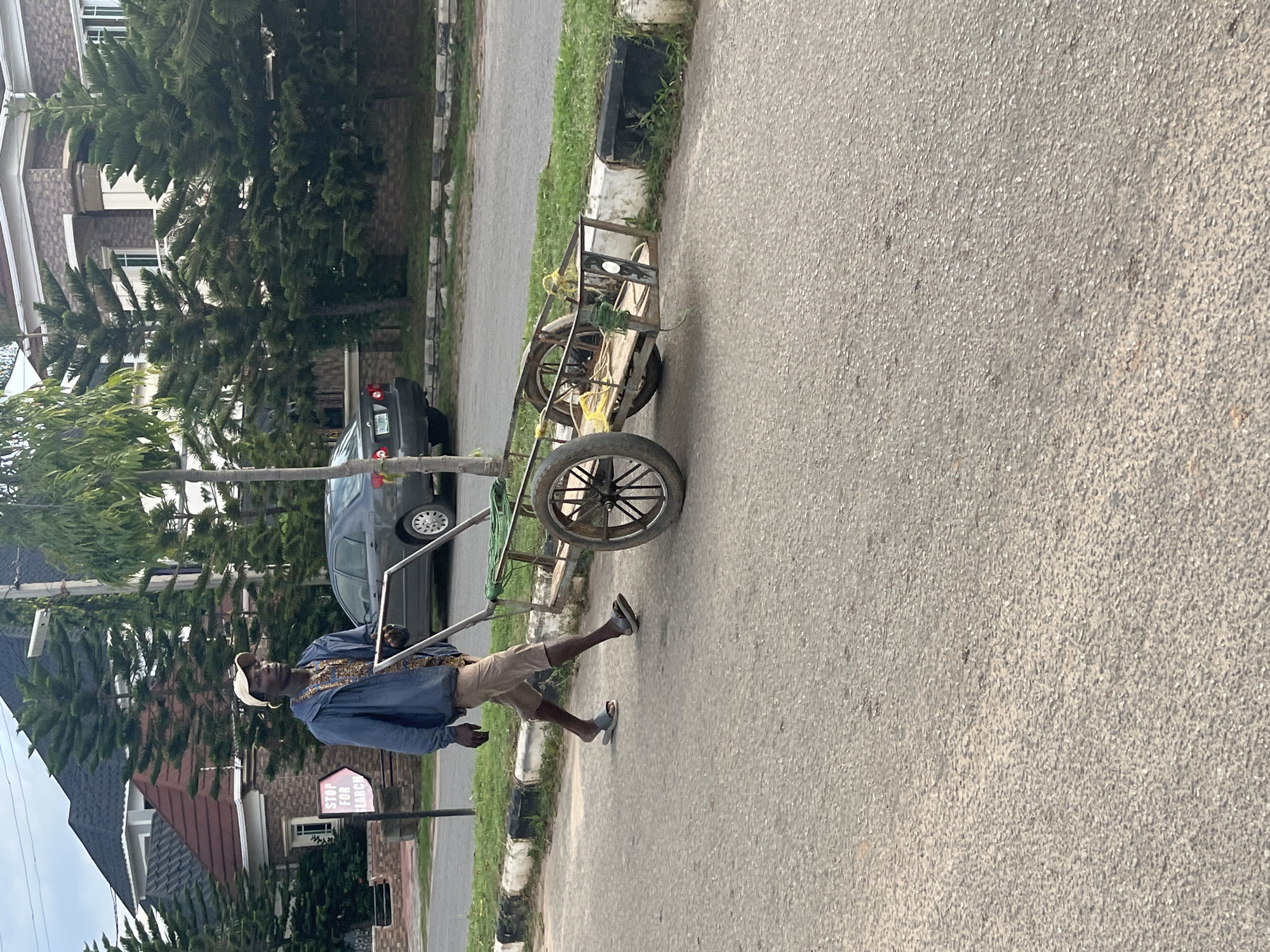 A dark-skinned adult male is depicted walking from left to right on an asphalt road, pushing a two-wheeled utility cart. He wears a beige baseball cap, a blue long-sleeved jacket over a patterned light brown shirt, khaki-colored shorts, and light grey slide sandals. His gaze is directed forward and slightly to the right.
The cart has a metal frame, wooden base, and two spoked wheels; it appears empty save for green and yellow ropes secured to its frame. The road surface is asphalt, flanked by a black and white painted concrete curb separating it from a grassy verge.
In the background, on the left, a white rectangular sign with red text reads "STOP FOR SEARCH" and features an arrow pointing to the right. Behind the man and extending to the right are several tall evergreen trees, likely Araucaria species. Further back, large, multi-story residential buildings with elaborate roofing (dark tiles and some terracotta) and light-colored facades are visible. A dark grey Toyota Corolla sedan is parked on the right side of the road, with a visible Nigerian license plate beginning with "ABJ," confirming the location in Abuja, Nigeria. The sky is overcast or hazy. The scene appears to be set during daytime, with soft ambient light.